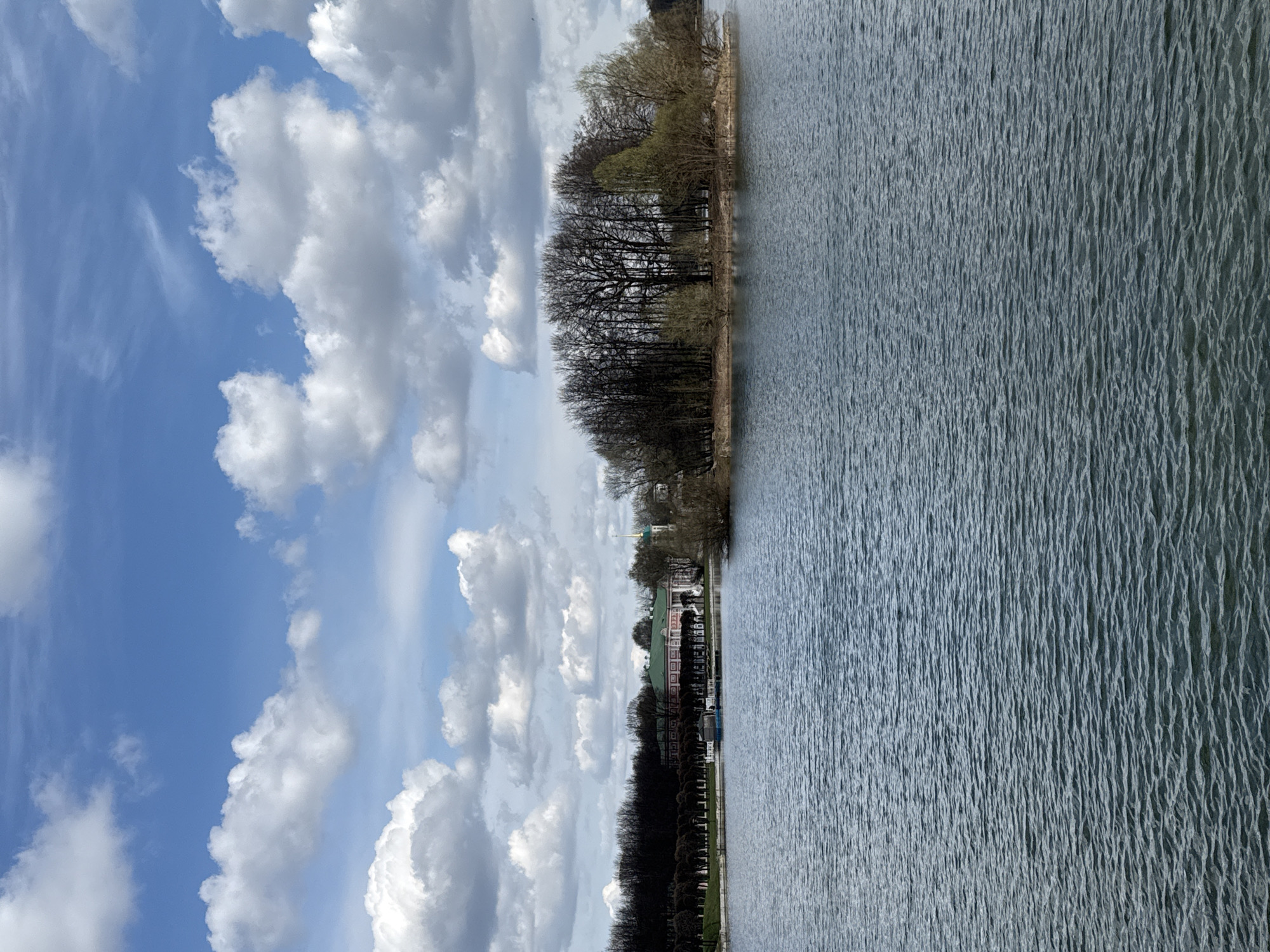 The image depicts a view of a body of water, possibly a lake or a wide river, adjacent to a park and buildings. The water surface is rippled, indicating a breeze. The sky is filled with fluffy white clouds against a blue backdrop. Along the shoreline, there are rows of trees and bushes forming a park. Buildings with a distinctive red and white facade are visible among the trees. The location is Moscow, Russia, specifically the Tsaritsyno Museum-Reserve, with the body of water being the Upper Tsaritsyno Pond. The scene appears peaceful and natural, with architectural elements blending into the landscape. No people are clearly visible.