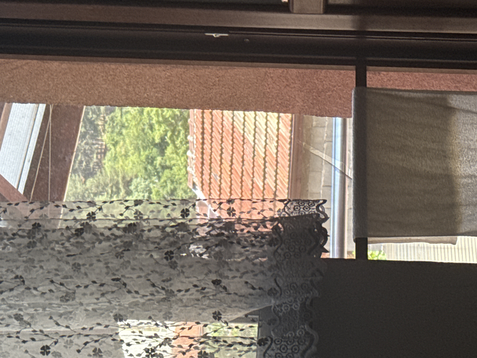 A close-up, eye-level shot captures a view through a window. In the foreground, a sheer, white lace curtain with a floral pattern drapes across the left side of the frame. Below the curtain, a thick, darker fabric hangs, obscuring the lower portion of the window. The middle ground reveals a residential scene outside: a section of an orange-tiled roof is visible, with a hint of a wall structure beneath it. Beyond that, lush green trees form a dense backdrop, suggesting a somewhat rural or suburban setting. The far right of the image shows a textured, salmon-colored wall and a dark vertical frame, likely part of the window or door structure. A light-colored towel is hung on a horizontal rod across the lower part of the window. The lighting suggests it is daytime, with sunlight illuminating the scene from the right. The image does not contain any people or indicate specific actions. The view is consistent with a residential setting in Dupnitsa, Bulgaria, possibly overlooking a garden or neighboring houses with abundant vegetation.