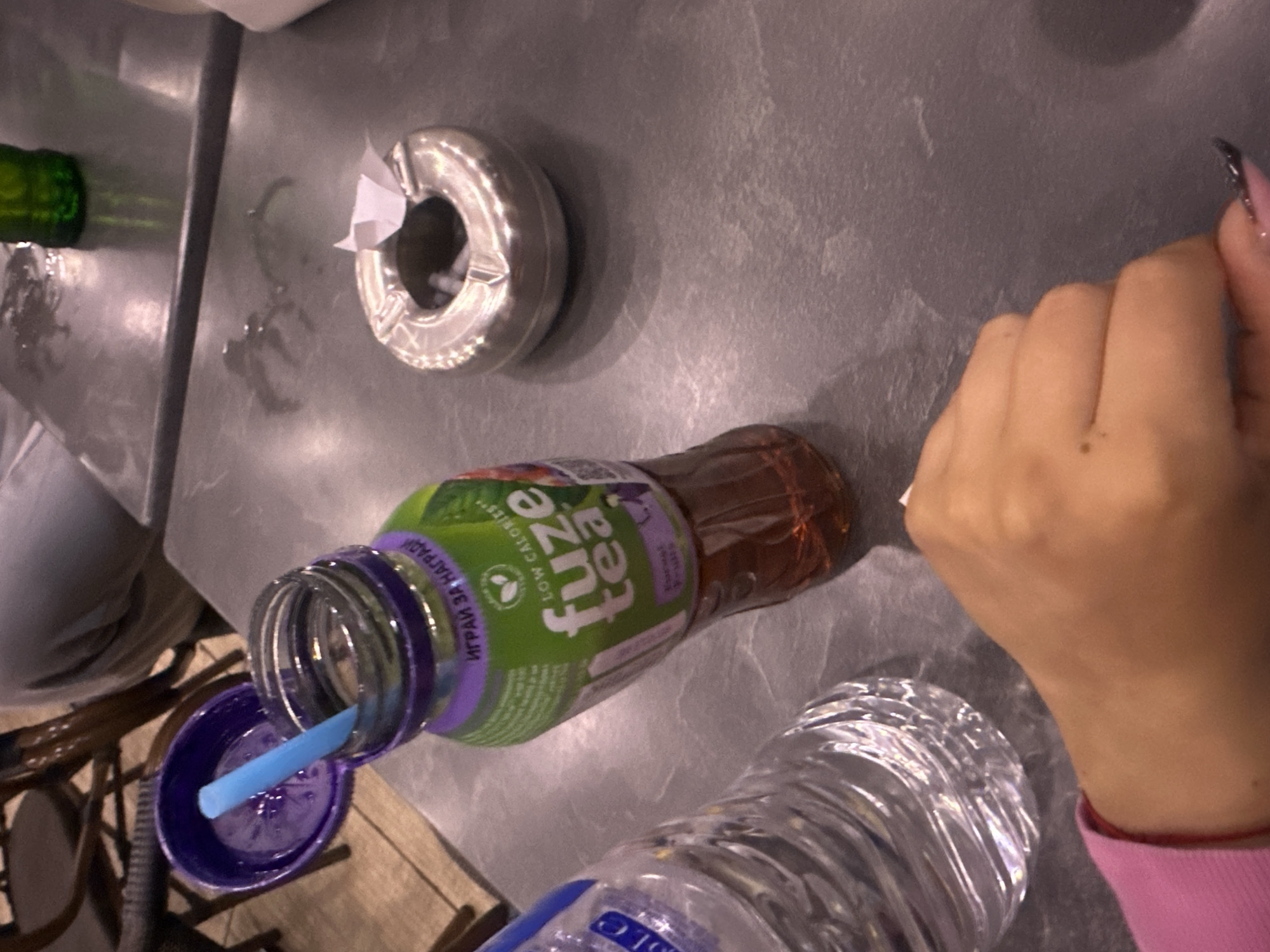 The image displays an overhead view of a table in an indoor setting, likely a cafe or a casual dining spot in Plovdiv, Bulgaria. The table surface is a dark gray or metallic color, showing some reflections and condensation marks.
In the center foreground, a plastic bottle of Fuze Tea is prominently featured, lying on its side. The bottle has a distinctive green and purple label, identifying it as "fuze tea" with "low calories" and a "peach" flavor. A light blue straw is inserted into the bottle's open neck, with the matching purple cap resting beside it. The label also displays text in Bulgarian, "ИГРАЙ ЗА НАГРАДИ," which translates to "Play for prizes." Next to the Fuze Tea bottle, a clear plastic water bottle is partially visible.
To the right, a person's hand is resting on the table, characterized by light skin tone and a dark-tipped, painted fingernail. In the upper part of the frame, a reflective, circular metallic object, possibly an ashtray or a napkin holder, is visible with a small piece of white paper inside. A portion of a green glass bottle is also present in the upper left corner, along with some moisture on the table. The background includes blurred elements of wooden furniture, suggesting chairs. The scene captures a casual moment of refreshment. The time of day is not explicitly indicated, and no specific emotions are discernible.