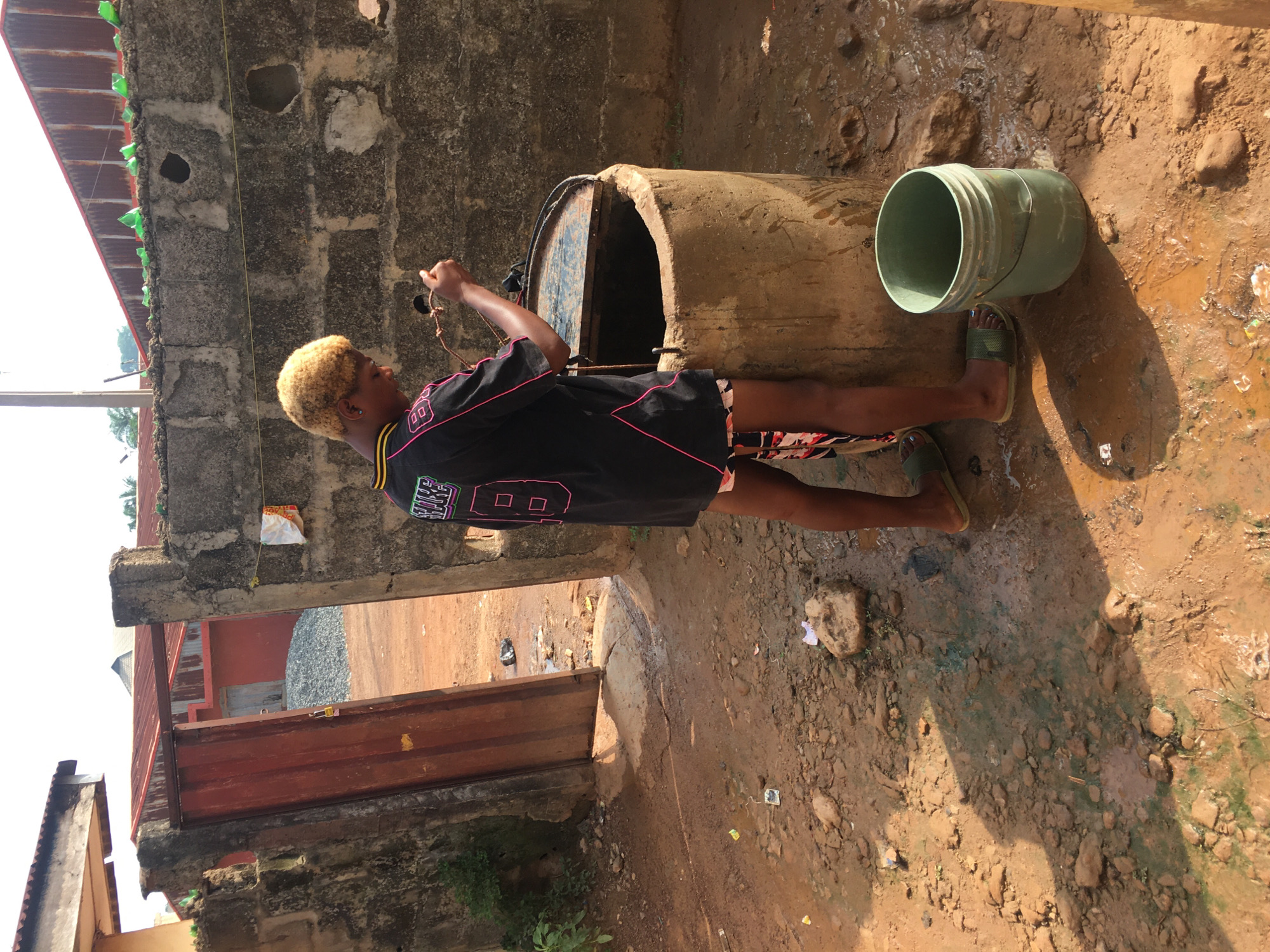The image captures an outdoor scene in Isieke, Nigeria, illuminated by bright daylight, casting strong shadows. A person with short, bleached blonde hair and dark skin is featured, dressed in a black t-shirt or jersey with pink trim, stylized graphics, and the number "00," along with patterned shorts and green slide sandals.
They are positioned beside a large, cylindrical, concrete-like structure, likely a well or water storage drum, and appear to be drawing water using a rope that extends into its dark opening. A light green plastic bucket lies on the reddish-brown, unpaved dirt ground nearby. The ground is uneven, littered with rocks and debris, and shows damp, muddy areas from spilled water.
The background consists of rough concrete block walls, some bare and some painted. A corrugated metal roof is visible, adorned with green plastic bottles along its edge, possibly for water collection or decoration. A rustic red metal gate or door can also be seen in the distance. The overall setting suggests a utilitarian area within a residential or communal environment.