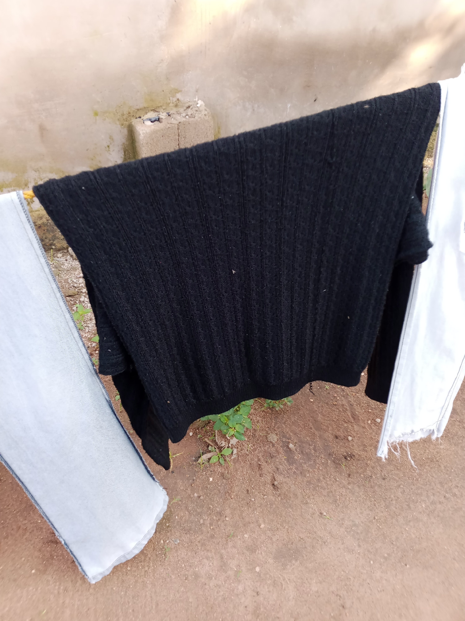 The image shows a black knitted sweater and a pair of light-colored jeans hanging on a clothesline. The clothes are outdoors, with a textured, light-colored wall in the background and a dirt ground below, sparsely covered with small green plants. To the left of the sweater, the leg of the jeans is visible, with frayed edges at the hem. To the right of the sweater, a piece of white fabric, possibly another garment, is also hanging on the line, with frayed edges. The scene suggests laundry is being air-dried. The lighting indicates it could be daytime, possibly under cloudy or diffused sunlight, given the absence of strong shadows. There is no visible text in the image. The setting appears to be a simple outdoor space in a residential area.