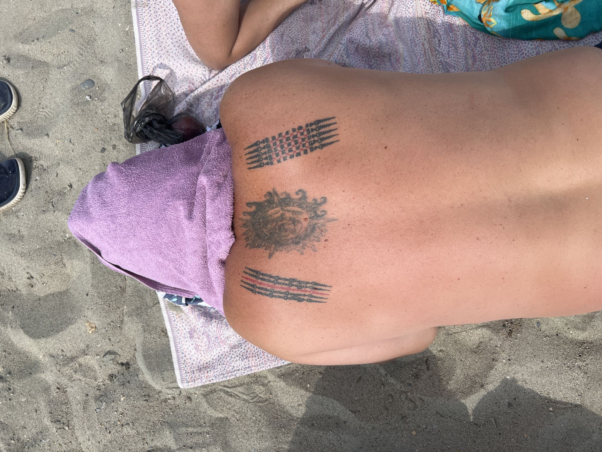 A person lies on a beach blanket on the sand, facing away from the camera. A purple towel is draped over their head. The person has tattoos on their upper back: a central tattoo of a face with radiating lines, and two patterned rectangular tattoos on either side. To the right of the person, a black plastic bag contains a red object. In the background, on the sand, are a pair of blue shoes. The image is taken outdoors on a sunny day.
