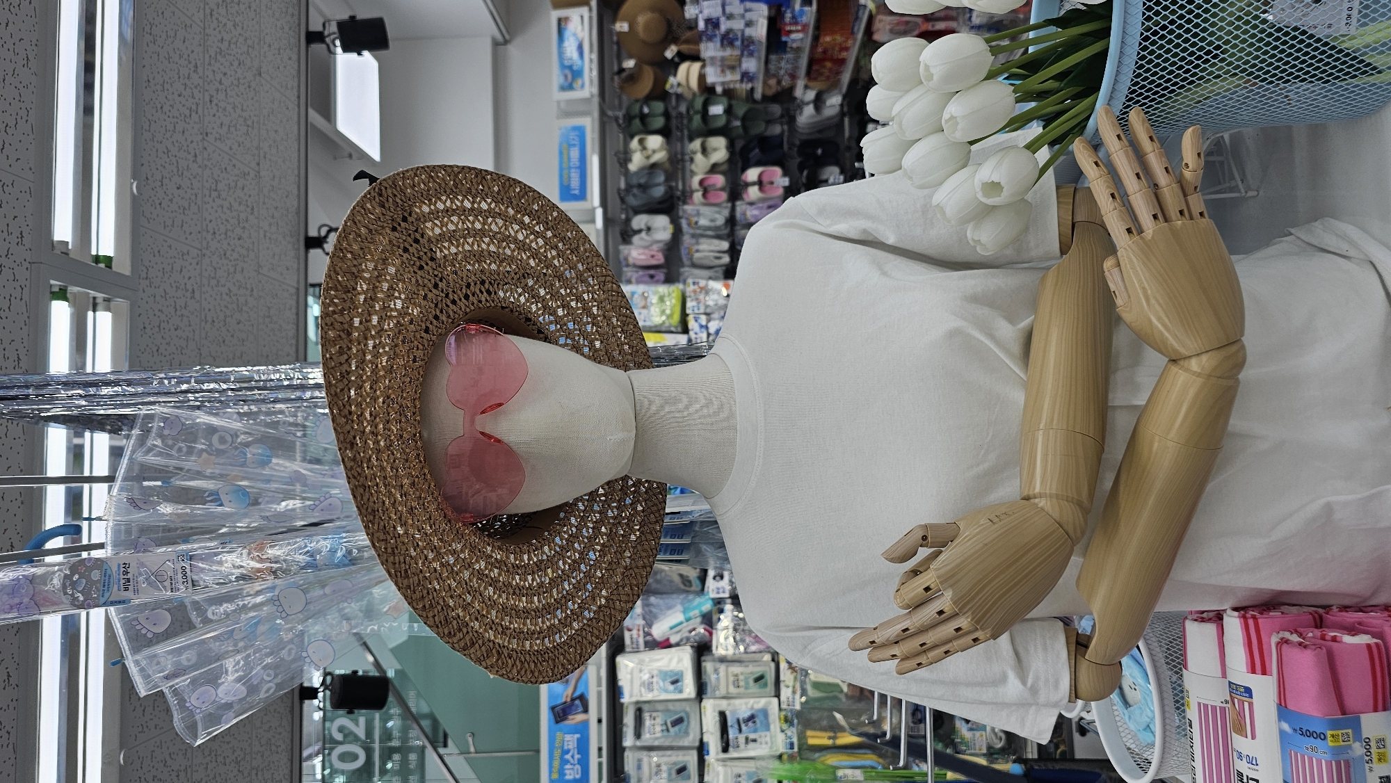 A mannequin wearing a straw hat with pink heart-shaped sunglasses and a white t-shirt is displayed in a retail store in Asan-si, South Korea. The mannequin's articulated wooden arms are positioned across its chest. In the background, shelves are stocked with various retail items, including umbrellas, toiletries, and footwear. To the right of the mannequin, a blue basket holds a bouquet of white tulips. The store appears to be well-lit, with fluorescent lighting fixtures visible on the ceiling.