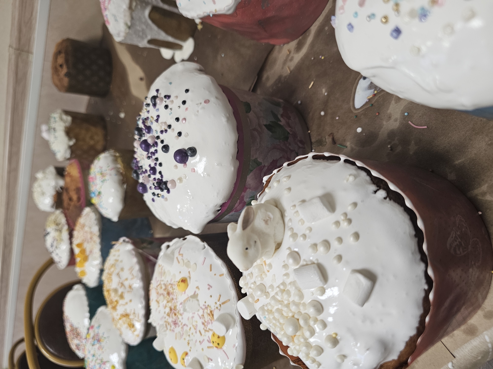 The image shows a collection of Easter cakes (Kulich) arranged on a brown surface, likely baking paper. They are decorated with white icing and various sprinkles, including small pearls, colored sugar strands, and marshmallows. One cake features a small white rabbit figurine. The cakes are of different sizes and shapes, some with floral designs on their paper molds. The environment seems to be an indoor setting, possibly a kitchen or a bakery. There is no clear evidence to determine the location being Moscow, Russia based on this image alone. No people are visible in the frame.