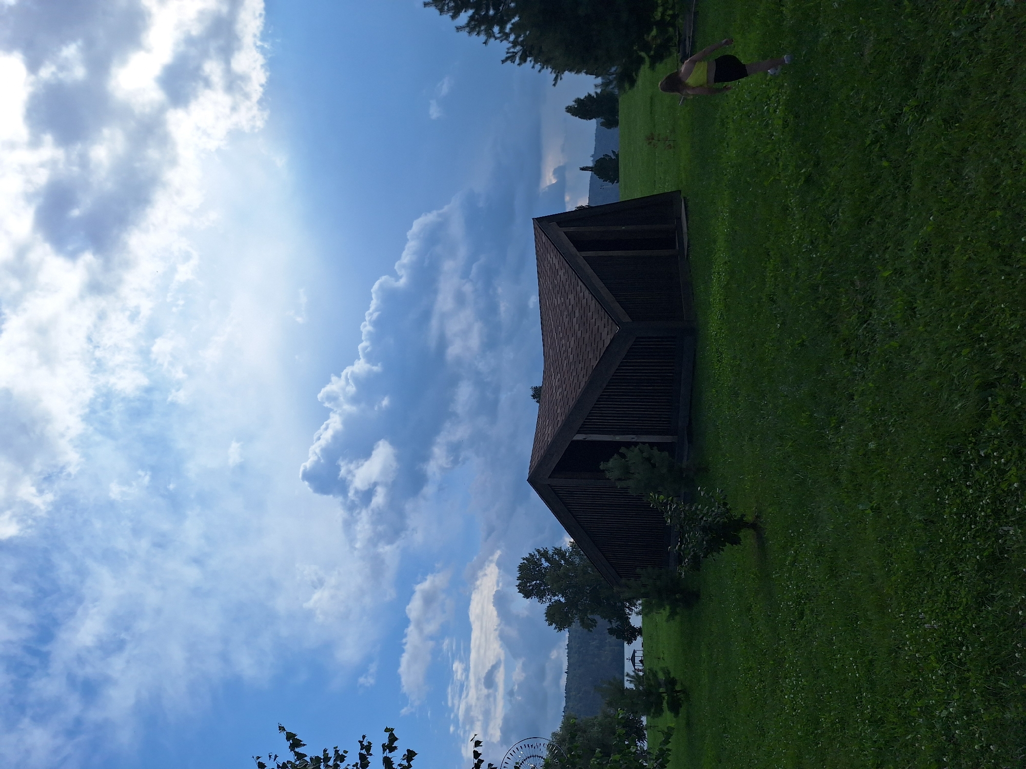 The image depicts an outdoor scene in Atamanovka, Russia, on a cloudy day. A dark, angular, wooden building with a pitched roof stands prominently in the mid-ground, situated on a grassy field. To the right of the building, a young person wearing a yellow top and black shorts is running away from the camera, their back to the viewer. The surrounding environment includes trees, a body of water in the distance with a small gazebo, and rolling hills or mountains on the horizon. The sky is a mix of blue and dramatic grey clouds, with some sunlight breaking through. The foreground is dominated by a lush green lawn with scattered white clover flowers.