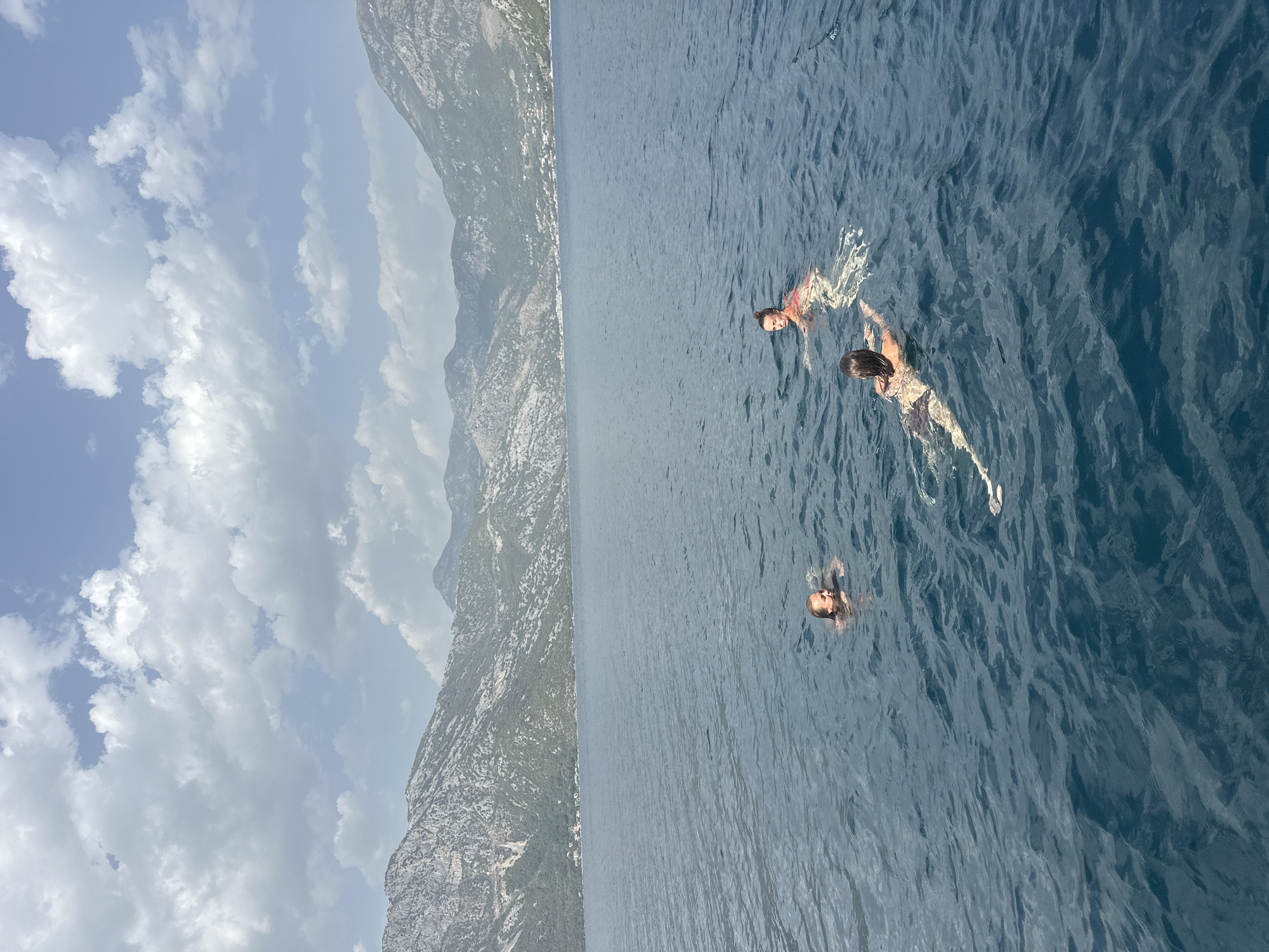 Three individuals are swimming in the blue, rippling waters of the Bay of Kotor, with the town of Perast, Montenegro, visible along the distant shoreline. The town is nestled at the base of steep, rocky mountains that dominate the background. The sky above is a mix of blue and white clouds.