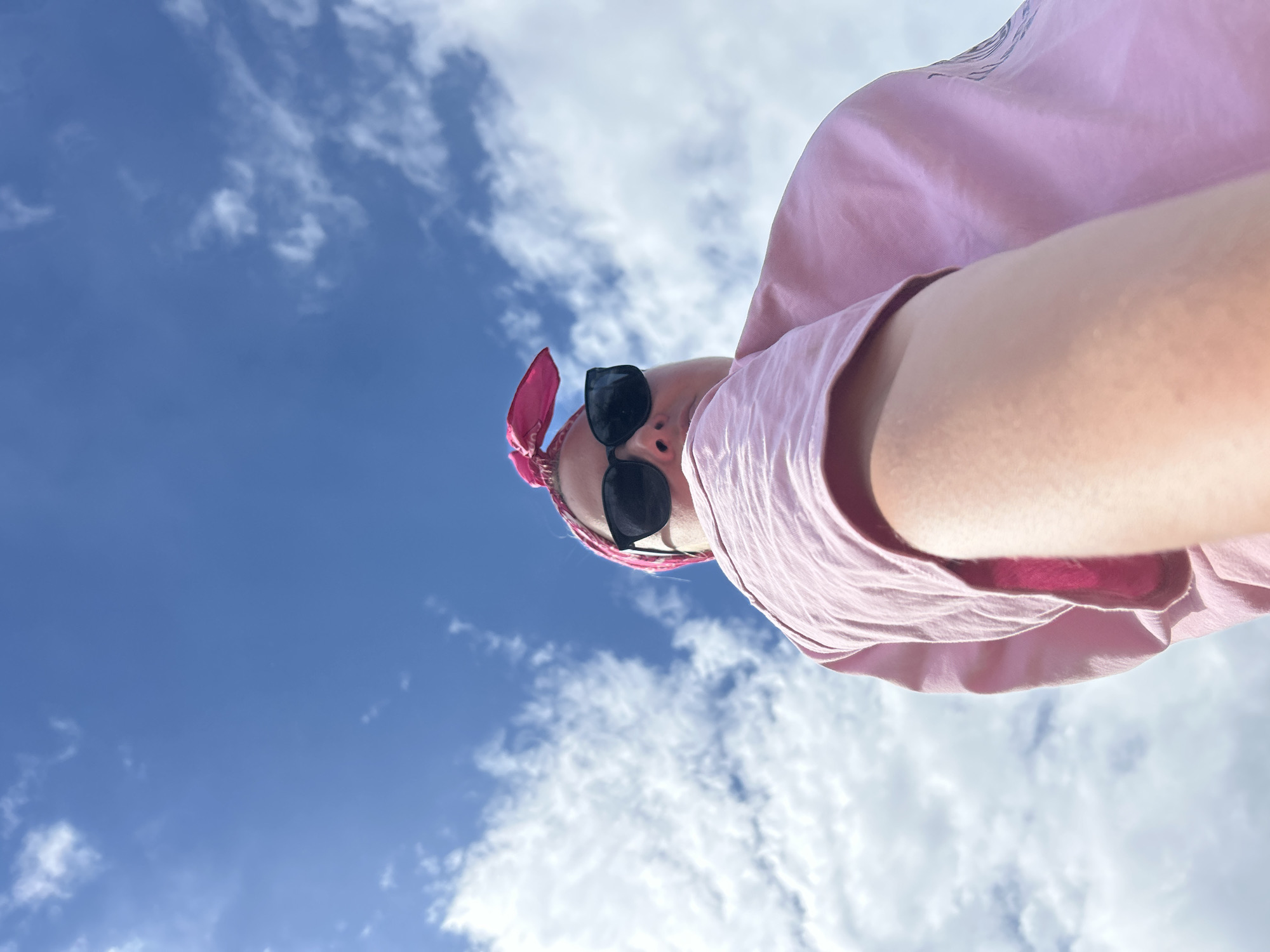 This is a low-angle, close-up, wide shot of a person outdoors against a bright blue sky with scattered white clouds, suggesting daytime and fair weather. The person is wearing a pink bandana tied around their head and black sunglasses. They are also wearing a pink t-shirt, and a portion of their arm is visible, indicating they are likely holding the camera to take a selfie or a photo from a low perspective. The camera's viewpoint is looking up towards the sky and the person, making them appear large in the frame. The overall impression is casual and summery. No text is visible in the image. Given the location context of Chaikovsky, Russia, this image could have been taken during the warmer months of the year.