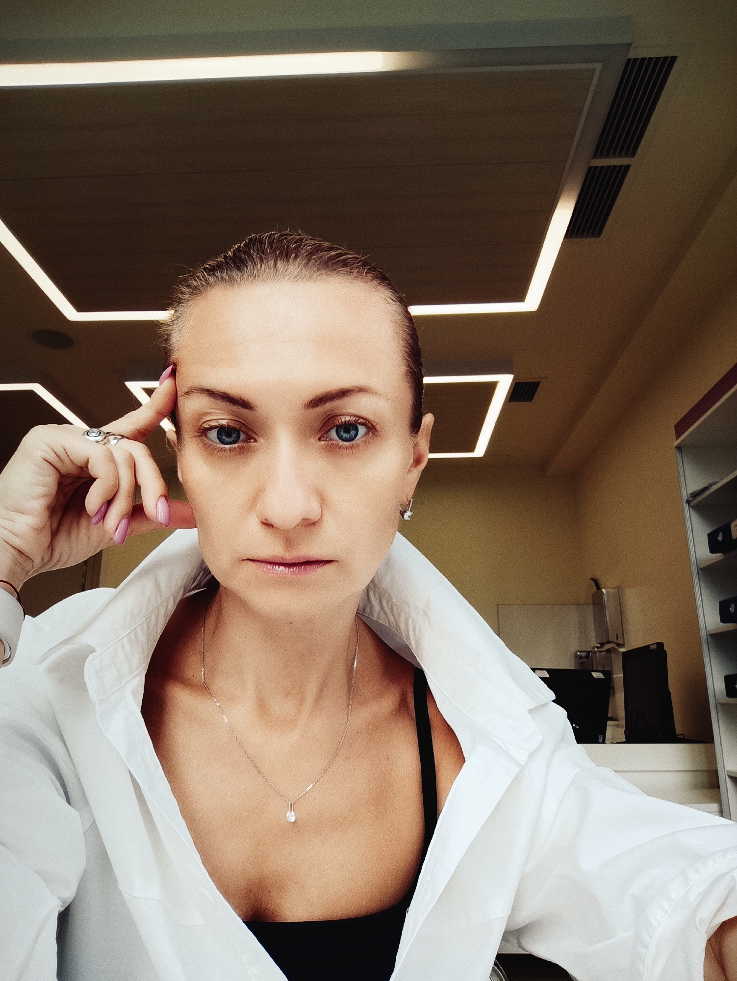 A woman with blonde hair pulled back and blue eyes is captured in a close-up selfie. She is wearing a white collared shirt, open to reveal a black bra and a delicate silver necklace with a pendant. Her left hand is raised to her forehead, adorned with rings on two fingers and a pink manicure. The background suggests an indoor environment with a tiled ceiling featuring geometric lighting fixtures and ventilation grates, hinting at a modern office or commercial space. The lighting is bright, casting a soft glow on her face and highlighting her features. The overall scene is a candid shot, possibly taken in Moscow, Russia, given the context provided.
