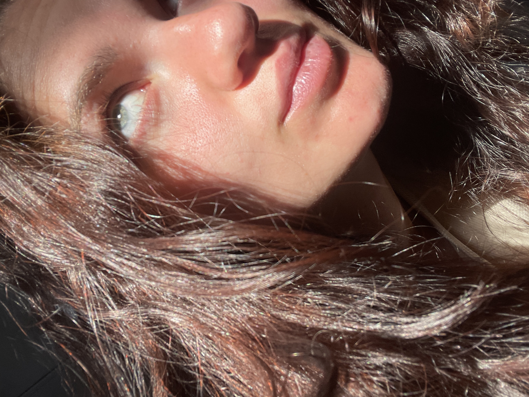 The image is a close-up, side-profile portrait of a young woman's face, partially obscured by her auburn hair. She has fair skin with warm undertones, illuminated by strong sunlight that casts highlights and shadows. Her light blue eyes are looking upwards and to the right, with visible eyelashes and eyebrows. Her lips are slightly parted, appearing moist and natural. The background is dark and out of focus, with some strands of her hair also in the foreground. There is no indication of the city of Мосметаллоконструкция Онт, Russia, or any other specific location within the image.