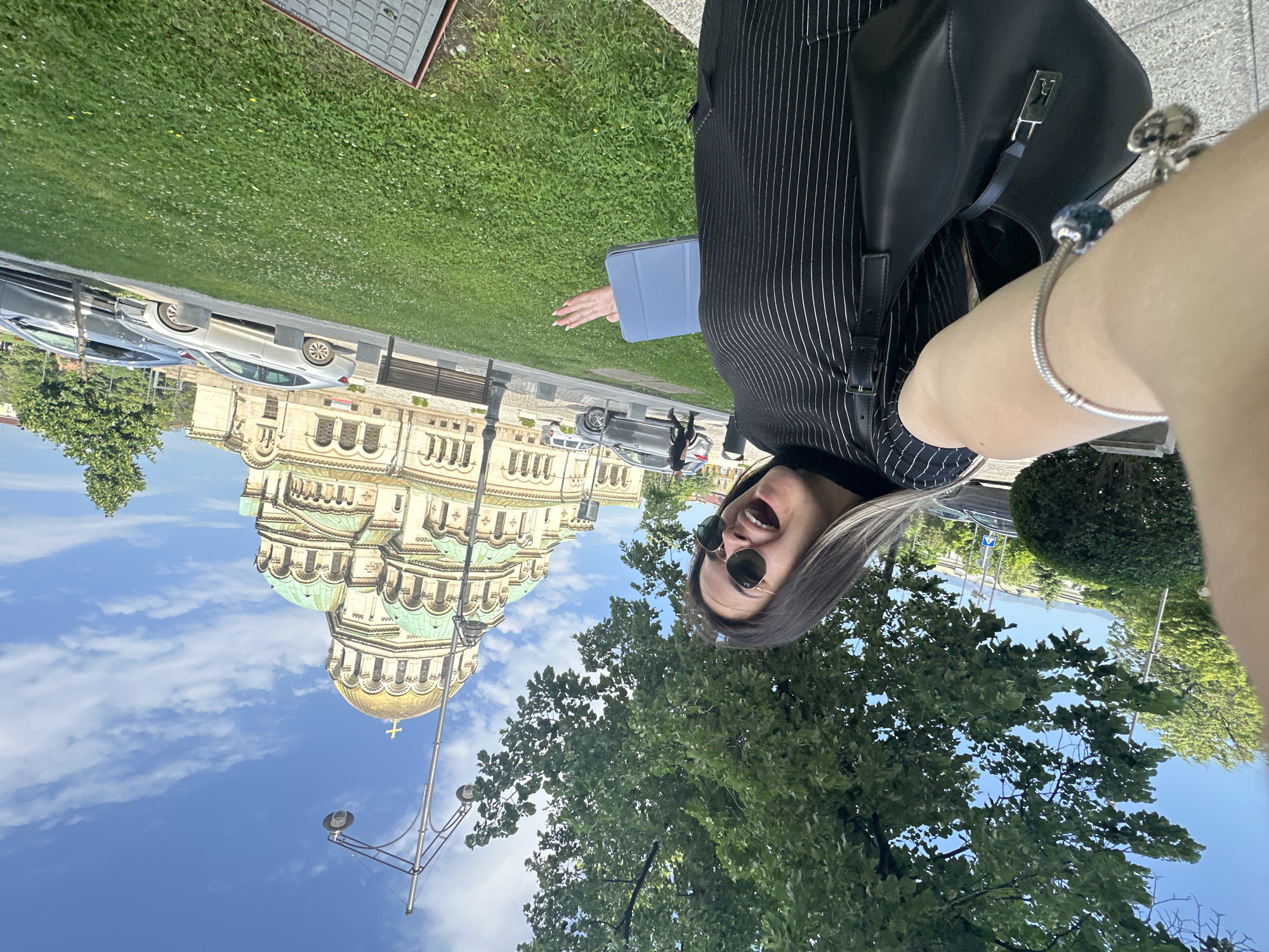 The image shows a woman taking a selfie in Sofia, Bulgaria. In the background is the Alexander Nevsky Cathedral. The woman is wearing sunglasses and a pinstripe jacket. She is holding a blue tablet and wearing a bracelet. Cars are parked along the street. The sky is blue with scattered clouds. A large tree is in the foreground.