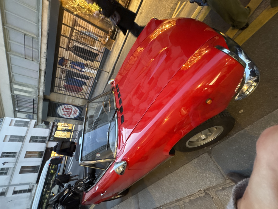 The image depicts a street scene at dusk or early evening on Rue Montmartre in the 1st arrondissement of Paris, France. The primary subject is a bright red, two-seater convertible sports car, resembling a Fiat 124 Spider, parked curb-side on a gray paved sidewalk. The car's soft-top is retracted, and its chrome front bumper and side mirrors are prominently visible.
In the mid-ground, across the street, are commercial buildings. A shop directly behind the red car features a closed metal security shutter adorned with painted illustrations of various menswear items on mannequins. A circular logo on this shutter prominently displays "ALO 1905". To the left of this shop, another establishment, partially visible, has a sign reading "E-CIGARETTES - E-LIQUIDES". Above these storefronts, a classic Parisian building with light-colored stone, multiple windows, and wrought-iron balconies extends upwards.
Several pedestrians are present. A person in a dark jacket and backpack walks away from the camera on the street behind the red car. Further back, near a bus stop, another individual in dark clothing walks leftward. A segment of a white city bus is visible moving in the background. On the far right of the frame, a blurry figure, likely a pedestrian, is moving past the car. In the extreme bottom-left corner, a portion of a foot and lower leg, wearing a dark shoe, belonging to the photographer, is visible. A black scooter or motorcycle is parked behind the red car on the sidewalk. Streetlights are illuminated, casting reflections on the wet-appearing street and the car's surface.