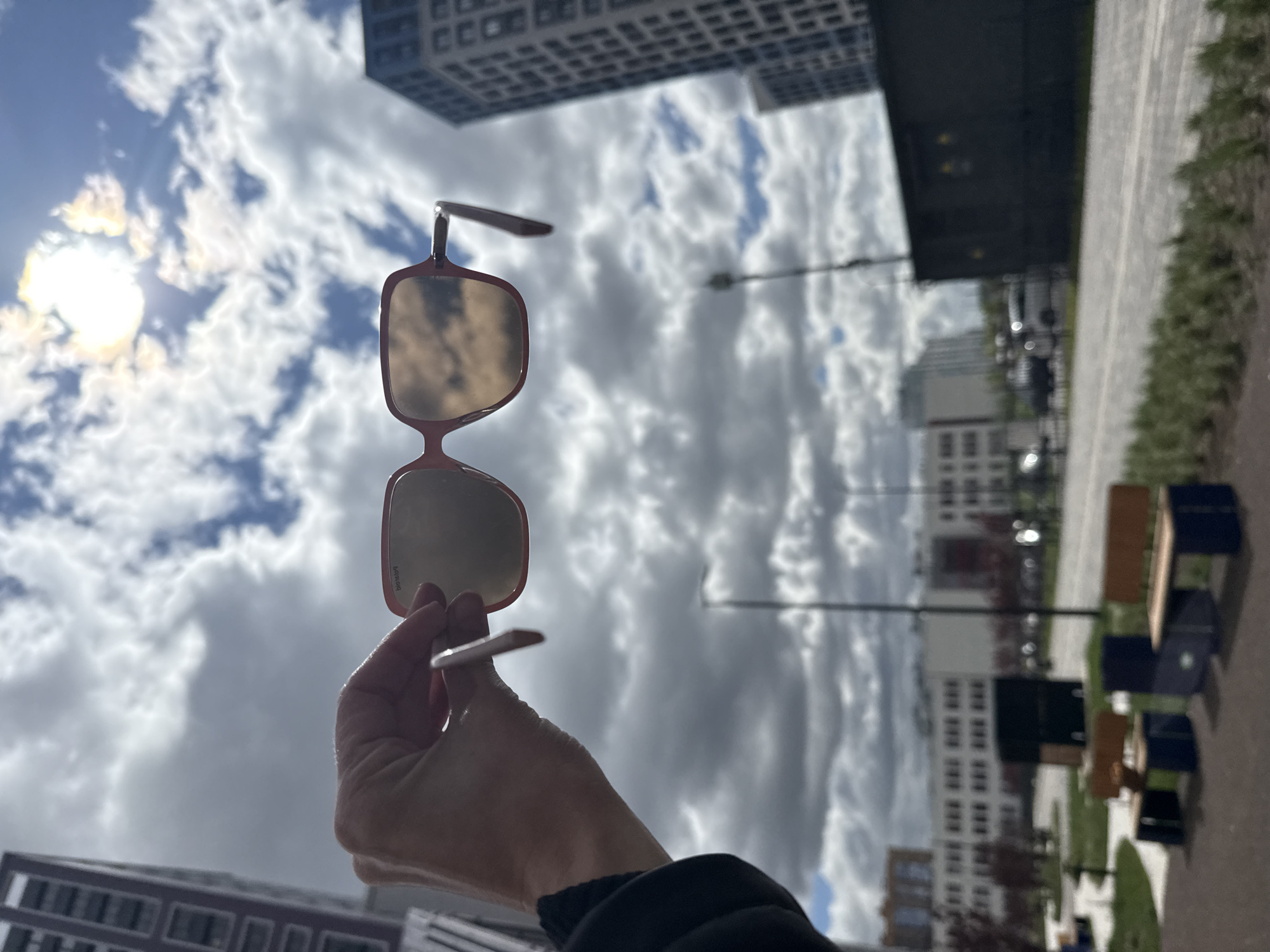 A hand holds a pair of pink-rimmed sunglasses against a backdrop of a cloudy sky and city buildings. The sunglasses reflect the surrounding environment, including the sky and buildings. The buildings are modern and appear to be residential or commercial. The location is likely in Saint Petersburg, Russia. A row of outdoor tables and chairs is visible on the right side of the image, adding to the urban setting.