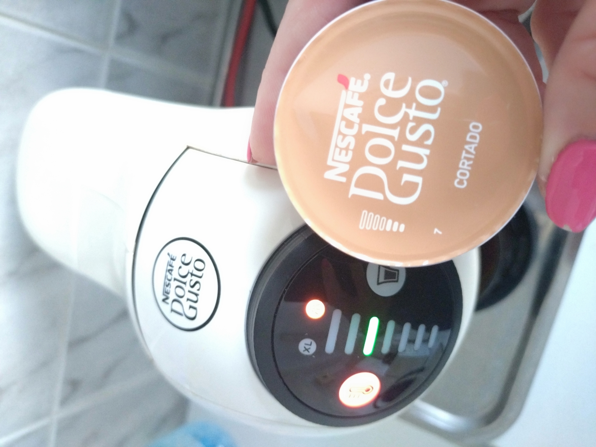 The image shows a white Nescafé Dolce Gusto coffee machine on a white tiled surface, presumably a kitchen countertop. A Nescafé Dolce Gusto "Cortado" capsule is held in someone's hand, ready to be inserted into the machine. The machine's control panel is visible, with several lights illuminated. The "XL" light is on, along with the power light. The lights indicate a selected coffee size. The location is unknown.