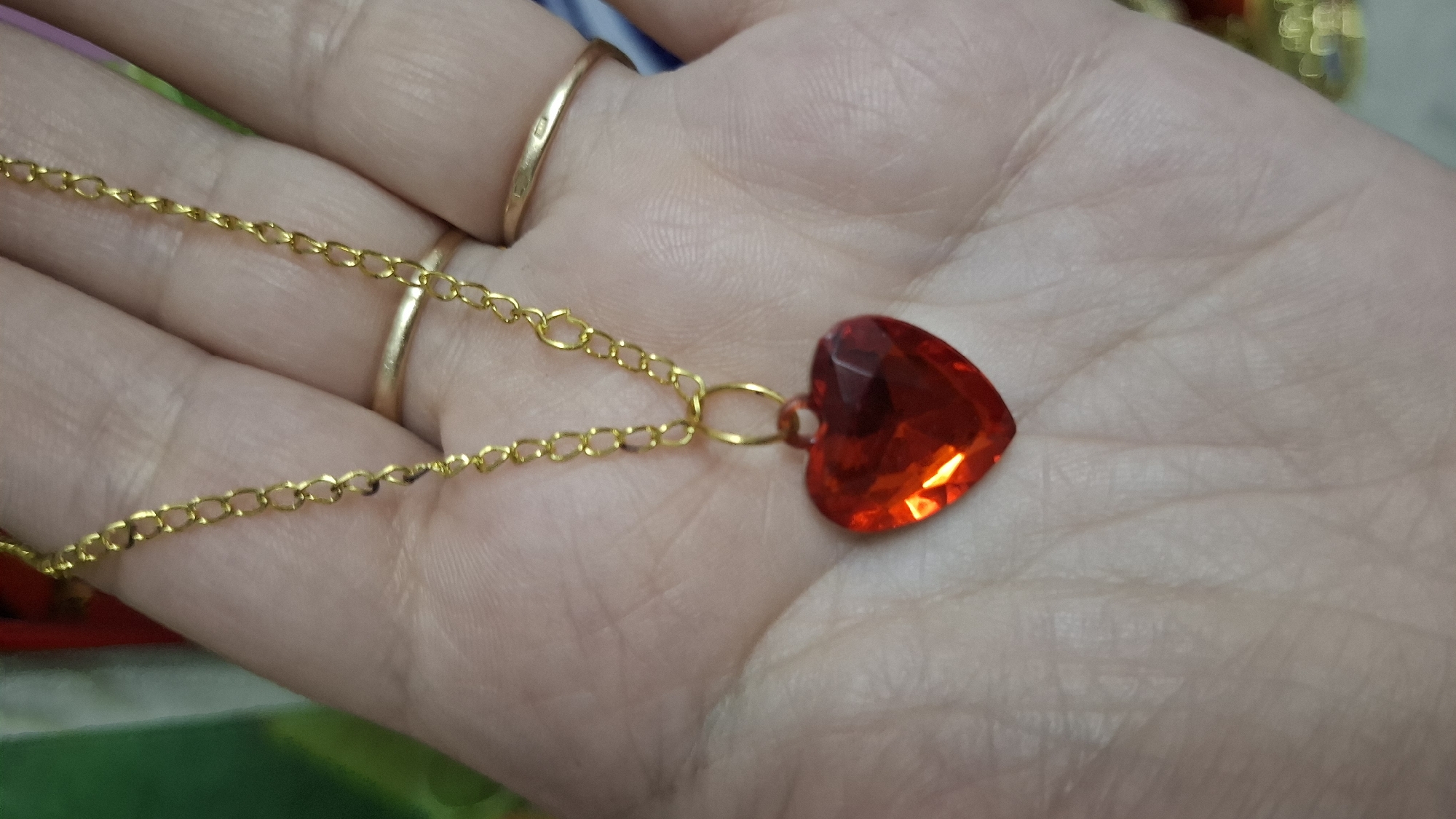 A close-up shot shows a person's palm with a gold chain necklace featuring a red heart-shaped pendant. Two gold rings are visible on the person's fingers. The background is blurred and appears to be indoors with some colorful elements. The location is not specified in the image.