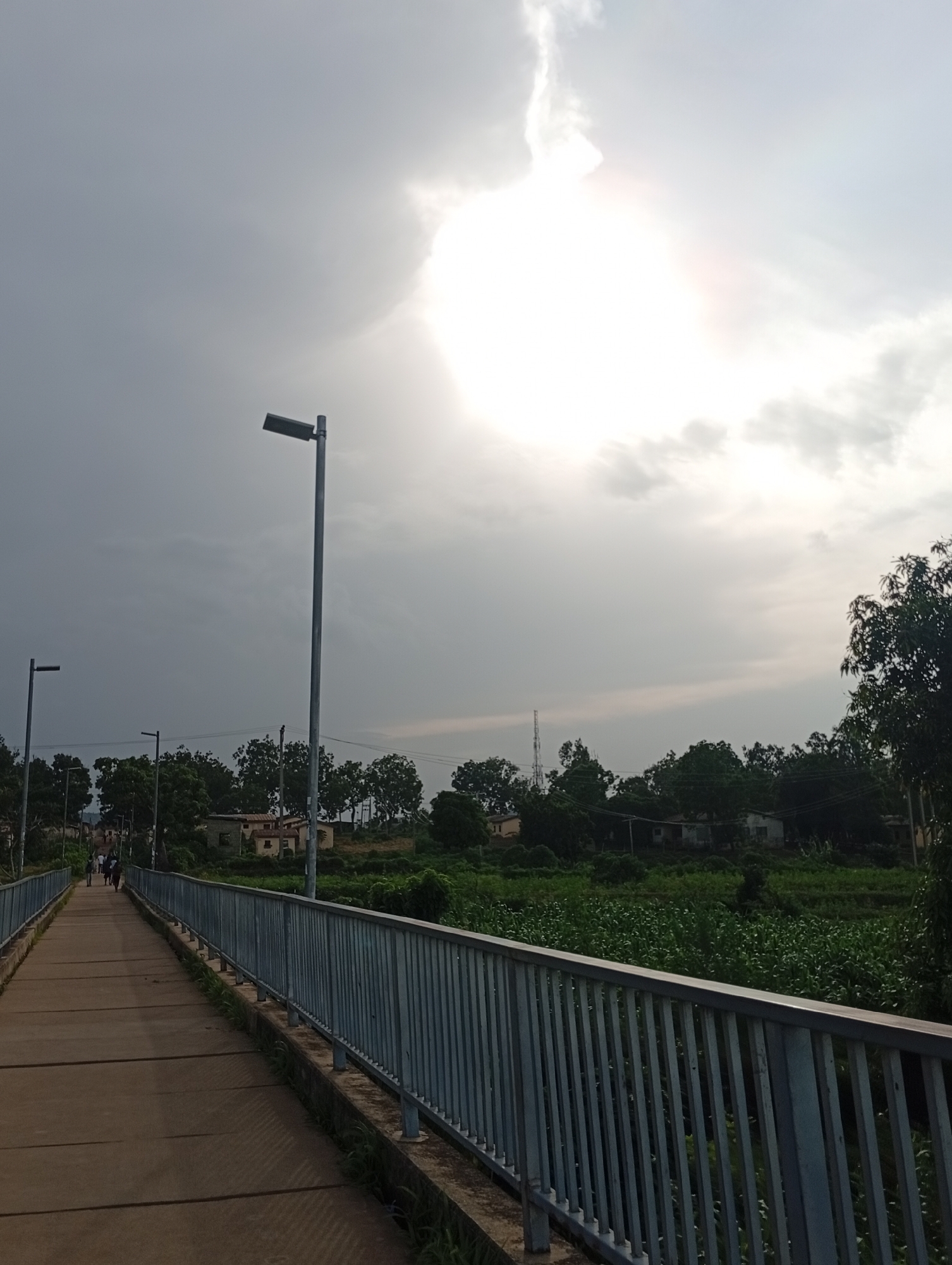 The image depicts a pathway or bridge with a metal railing on the right side, leading towards buildings and a vegetated area. The pathway appears to be made of concrete slabs with some grass growing between them. Several streetlights are visible along the pathway. In the background, there are trees, some low-rise buildings, and what appears to be agricultural fields. The sky is overcast with a bright sun shining through the clouds, creating a diffused light. A few individuals are walking on the pathway in the distance. The overall scene suggests an outdoor environment in a semi-urban or rural setting. The presence of streetlights and buildings indicates some level of development.