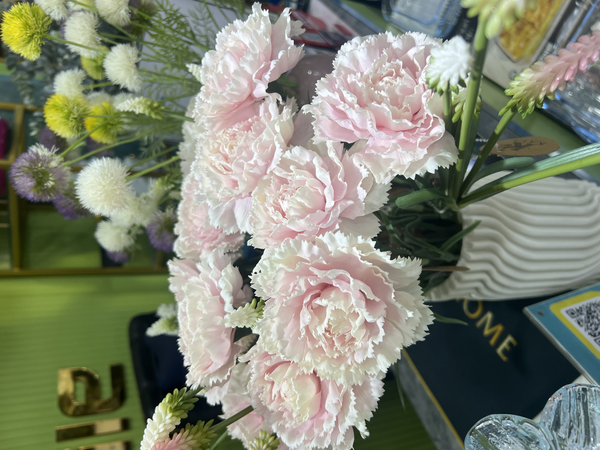 A close-up, slightly overhead shot captures a bouquet of soft pink carnations in a white ribbed vase. The carnations are the primary focus, with their delicate, ruffled petals filling the foreground. Behind them, various other artificial flowers are visible, including white pom-pom-like blooms, yellow spheres, and purple globe-shaped flowers, suggesting a floral arrangement in a shop or home. The background is out of focus, but a vibrant green wall with metallic letters that appear to spell "ALE" is discernable, along with other shelves and objects that hint at a commercial setting. The overall impression is one of artificial floral beauty in a brightly lit indoor space. This image does not contain enough information to determine its location in Eshanguzar, Uzbekistan.