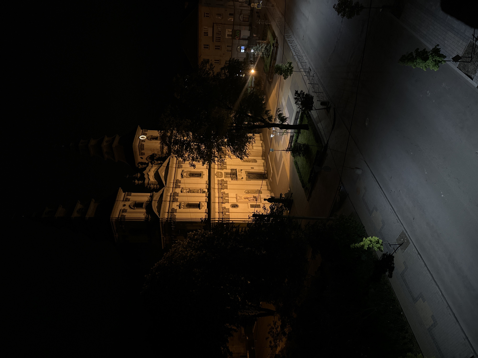 The image depicts a nighttime view of the Church of the Exaltation of the Holy Cross in Ternopil, Ukraine. The church, with its visible towers and ornate facade, is illuminated by artificial lighting. Trees are present in the foreground and around the church. A street runs along the side of the church with a park area. A building is visible in the background of the photo.