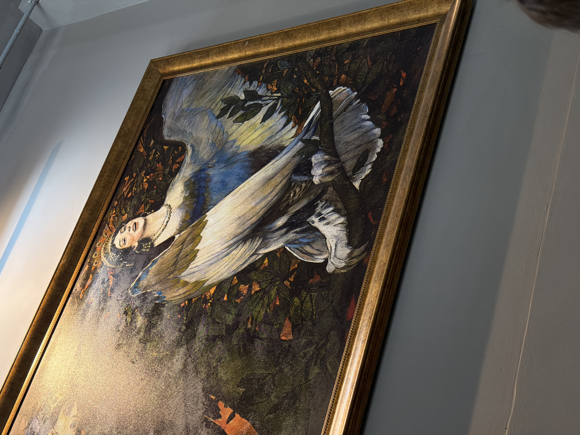 The image shows a framed painting of a mythical winged creature with a human woman's head. The creature has large, feathered wings and is perched on a branch amidst foliage. The woman has dark hair, is wearing jewelry, and has an expressive face. The painting's style appears to be classic or Art Nouveau. The painting is hanging on a wall in a public space. No specific location in Đà Nẵng, Vietnam is identifiable from the image.