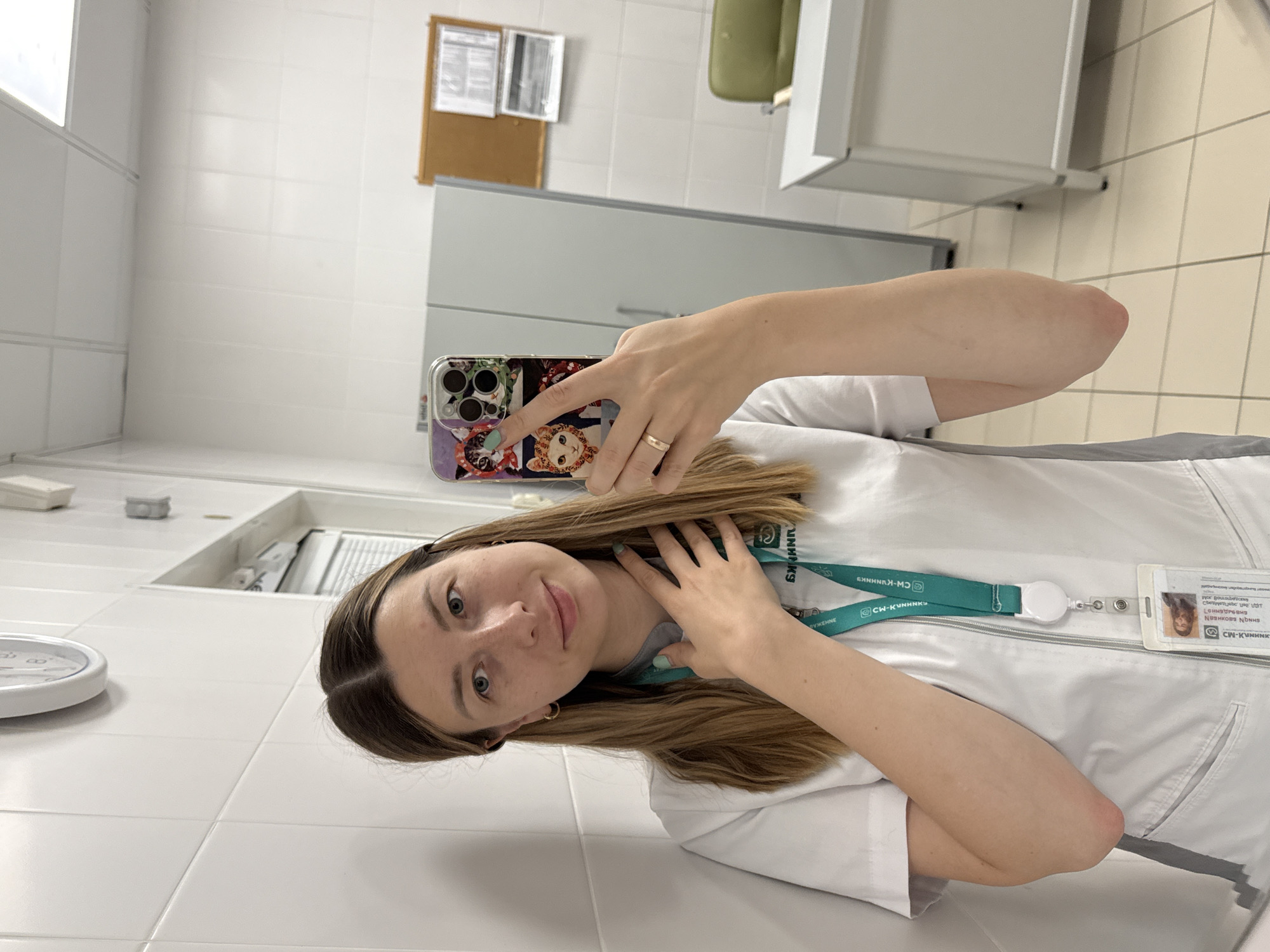 A young woman is taking a mirror selfie in what appears to be a medical or laboratory environment. She has long, light brown hair, blue-grey eyes, and a pleasant, calm expression with a slight smile. She is wearing a white medical uniform or lab coat and a green lanyard around her neck, holding an ID badge. On her right hand, she wears a simple gold ring and holds a smartphone with a decorative case featuring cartoon cat-like characters.
The setting is an indoor, brightly lit room with white tiled walls. In the background, there is a light-colored counter, a white cabinet, and a cork board with papers pinned to it. A round white object, possibly a clock or timer, is visible on the counter to the left of the woman.
The green lanyard prominently displays Cyrillic text, including "СМ-Клиника" (SM-Clinic), a common network of medical centers, consistent with the provided location context of Moscow, Russia. Her ID badge, also attached to the lanyard, shows her photograph and further Cyrillic text, much of which is fragmented but reinforces the medical facility context. Her left hand is gently placed on her chest, near her collarbone.