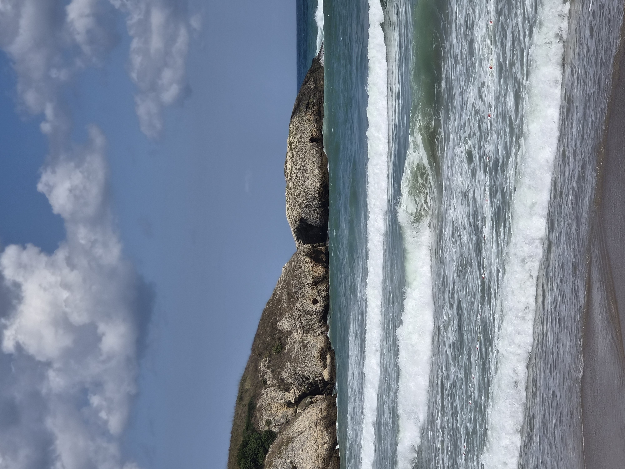 The image depicts a scenic view of a coastline in Şile, Turkey, featuring a rocky cliff face on the left and a larger rock formation in the sea on the right. The water is a vibrant turquoise, with white waves crashing onto the shore. The sky is a clear blue with scattered white clouds. A small, dark sand beach is visible in the foreground. There are no people or visible man-made structures, suggesting a natural, uninhabited landscape. The scene is captured in a vertical orientation, emphasizing the height of the cliffs and the expanse of the sea.
