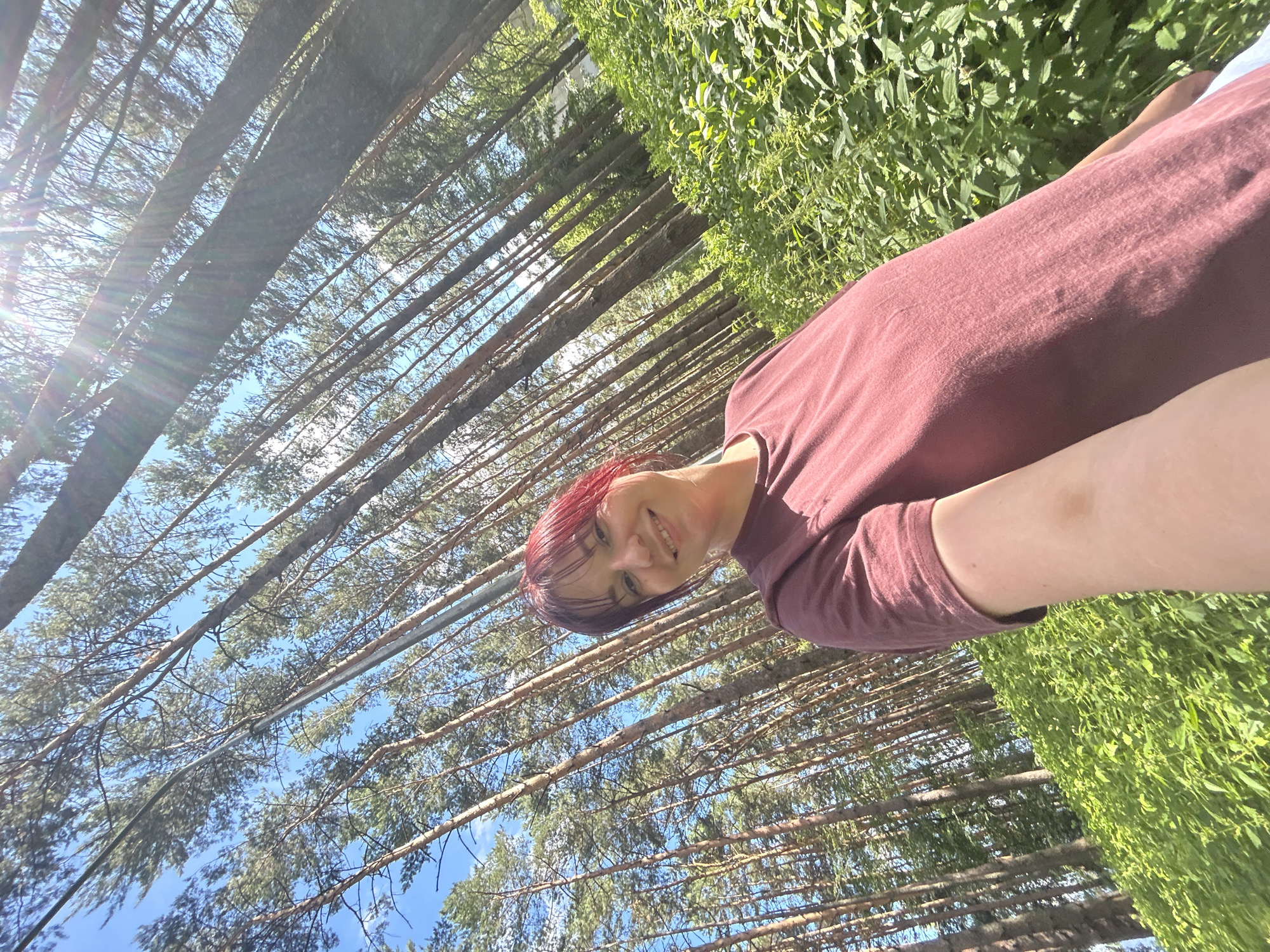 The media file is a selfie of a person with red hair, smiling. They are wearing a maroon t-shirt. The background is a dense forest with tall, thin trees and bright green foliage. The sun is shining brightly, casting lens flare across the image and illuminating the sky. The perspective is from below, looking up through the trees. The overall impression is a cheerful and sunny day spent outdoors in a natural setting. The image is oriented vertically. There is no visible text. The activity is a person taking a selfie in a forest. The time of day appears to be daytime, likely sunny and warm given the brightness and lens flare. The location cues suggest a natural environment, possibly a park or wooded area. The emotion displayed is happiness.