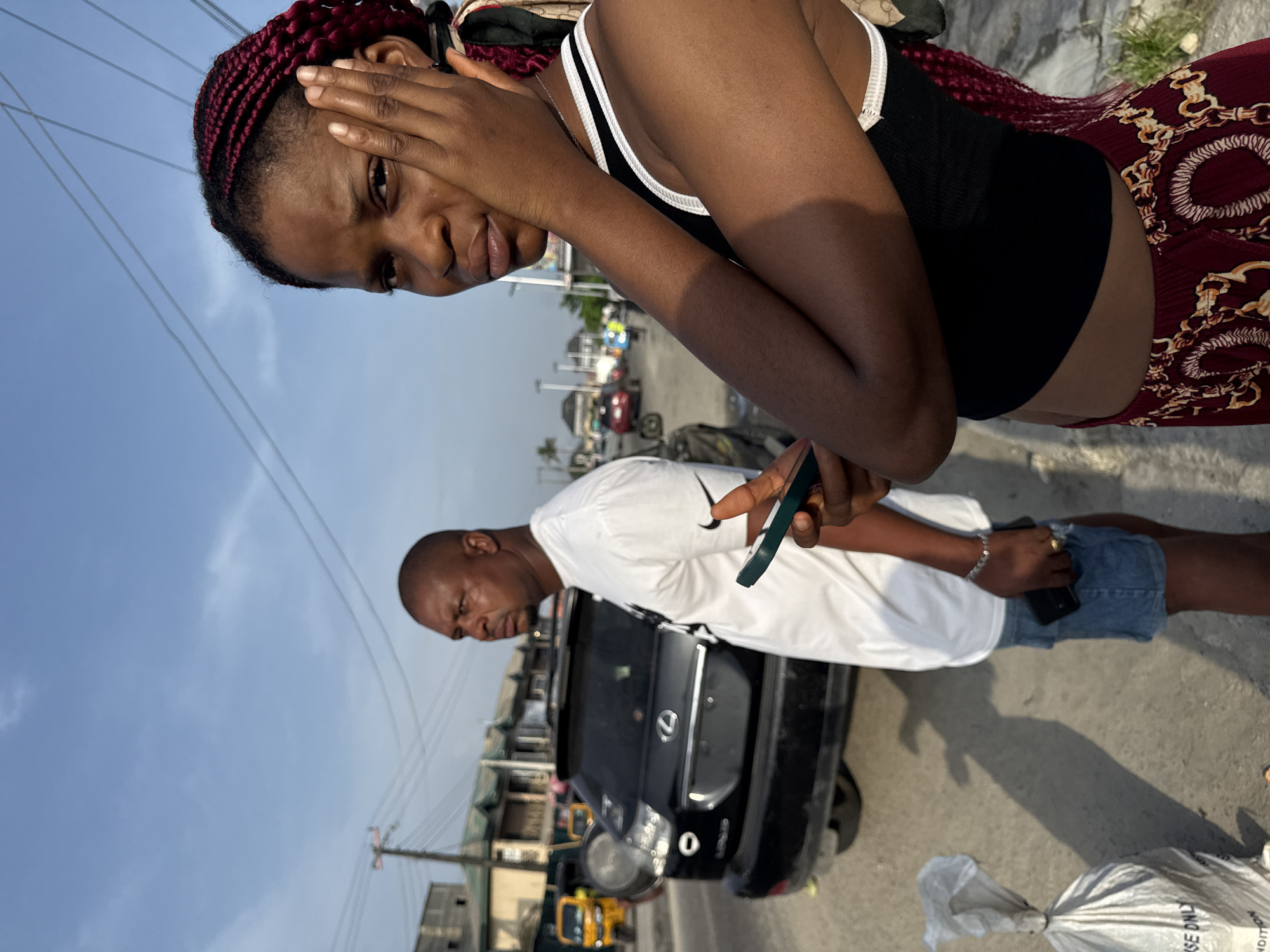 The image shows a woman with red braided hair, wearing a black tank top with white trim, standing next to a man in a white Nike t-shirt and denim shorts. She has her hand on her forehead and appears distressed. The man is holding a phone. In the background is a black car, possibly a Lexus, and a street scene with buildings, a yellow taxi, and other vehicles. The location is possibly in Ado, Nigeria, given the appearance of the vehicles, buildings, and overall street scene.