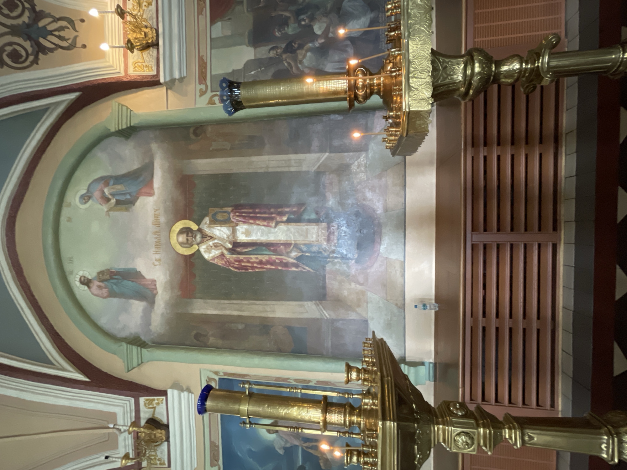 The image captures an interior view of a Russian Orthodox church, specifically within the Trinity Lavra of St. Sergius in Sergiev Posad, Russia. The focal point is a large mural depicting a saint, likely Saint Nicholas, with a halo, wearing ornate religious vestments. He stands centrally, holding a book. Above him, to the left, is a depiction of Jesus Christ, and to the right, the Virgin Mary, both with halos and depicted as if on clouds. Flanking this central mural are other religious paintings. In the foreground, two large, ornate brass candelabra with lit candles stand prominently. One candelabrum is on the left, and the other is on the right, partially obscuring the background. The walls are painted with decorative arches and molding. Below the murals, there is a wooden structure with slatted panels, possibly a cabinet or a radiator cover. A plastic water bottle is visible on the wooden surface. The floor appears to be tiled in a geometric pattern. The overall lighting suggests an indoor setting, with light coming from the candles and ambient church lighting.