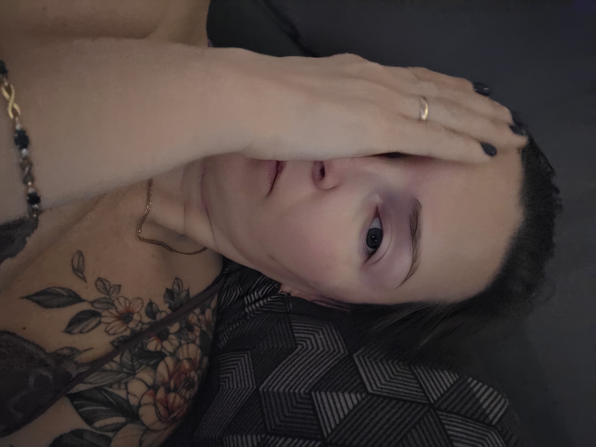 A woman is lying down, facing the camera. Her right hand is covering the upper part of her face, obscuring her forehead and eyebrow. She has a gold ring on her hand. Her left eye is visible and open. She has a gold necklace and a bracelet on her left wrist. A floral tattoo is visible on her upper chest. She is lying on a pillow or cushion with a geometric pattern, and the background appears to be a dark surface, possibly a sofa or bed. No specific details indicate this image was taken in Chaikovsky, Russia.