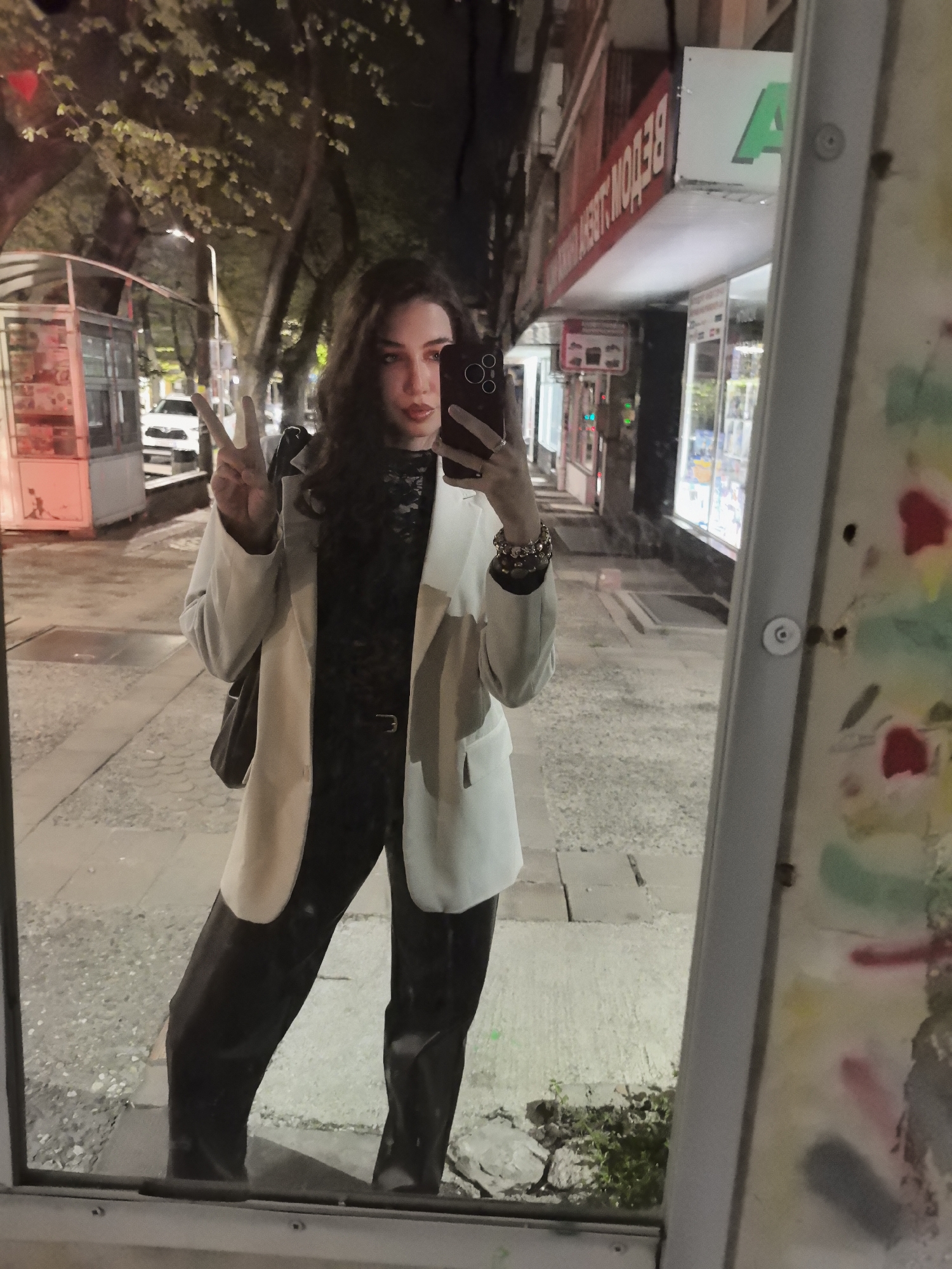 A young woman takes a mirror selfie on a city street at night. She wears a white blazer, a dark patterned top, black pants, and several bracelets. She is holding up a phone to take the picture, and making a peace sign with her left hand. The street behind her includes a small kiosk, a sidewalk, buildings with lit storefronts, and trees. The storefront signs are written in Cyrillic, identifying the location as Stara Zagora, Bulgaria.