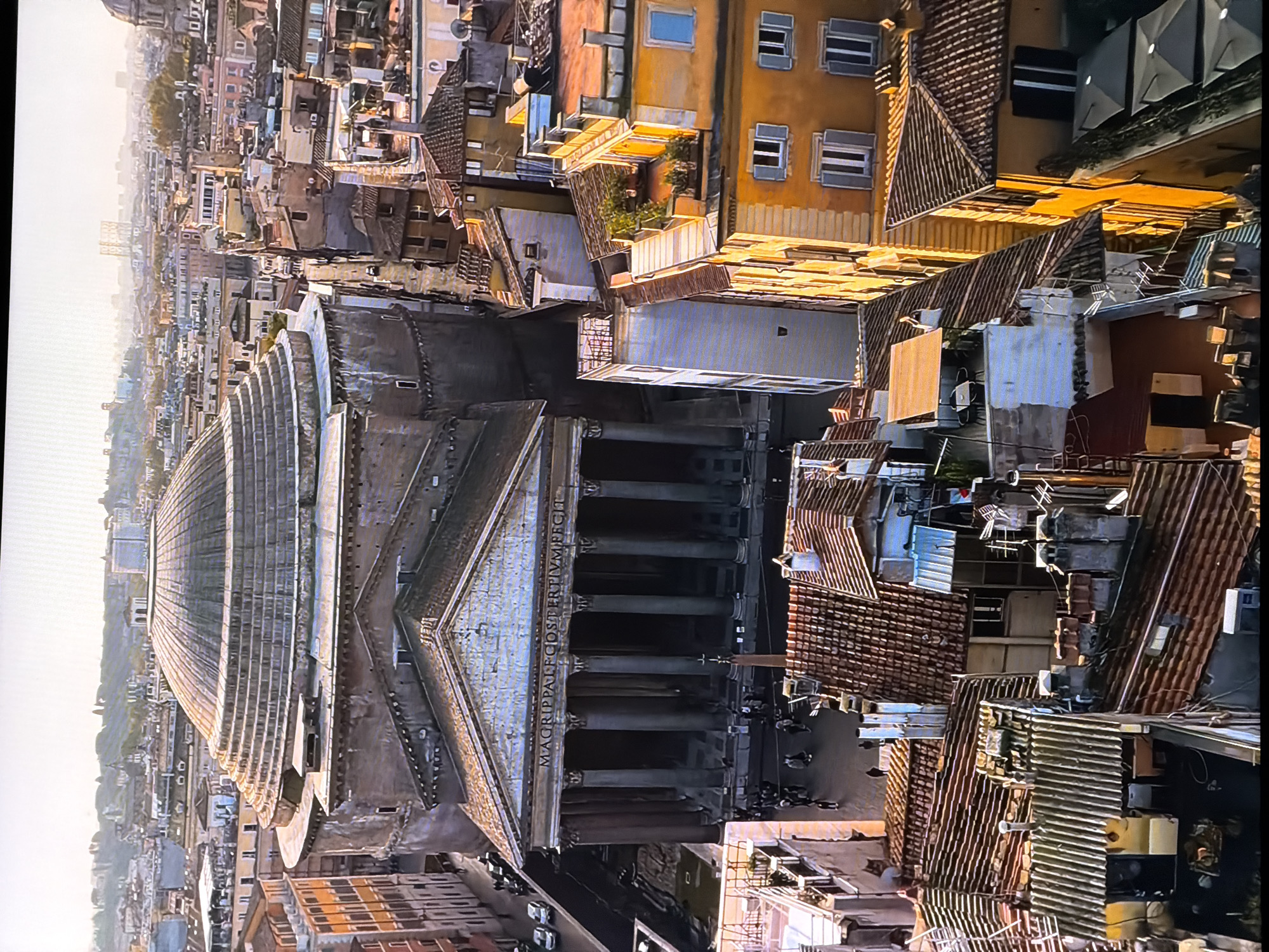 This is an aerial, slightly angled, full-shot image of the Pantheon in Rome, Italy. The Pantheon is a massive, ancient Roman temple, now a church, with a prominent dome and a colonnaded portico. The image captures the surrounding urban landscape, characterized by dense, multi-story buildings with terracotta tile roofs, typical of European cities. The buildings are of varying architectural styles and colors, with some exhibiting classical features and others appearing more modern. The sunlight casts long shadows, indicating it is likely late afternoon or early morning. Several cars are visible on the street in front of the Pantheon, and small groups of people are scattered around the plaza. The sky is clear and bright. The city of Chirchik, Uzbekistan is not depicted in this image.