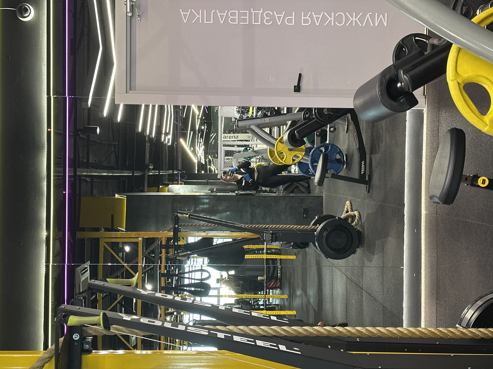 A gym interior in Perm, Russia, features exercise equipment. A woman is visible on a weight machine. Signage in Cyrillic script is present. The gym is brightly lit with neon lighting. Ropes are part of the equipment.
