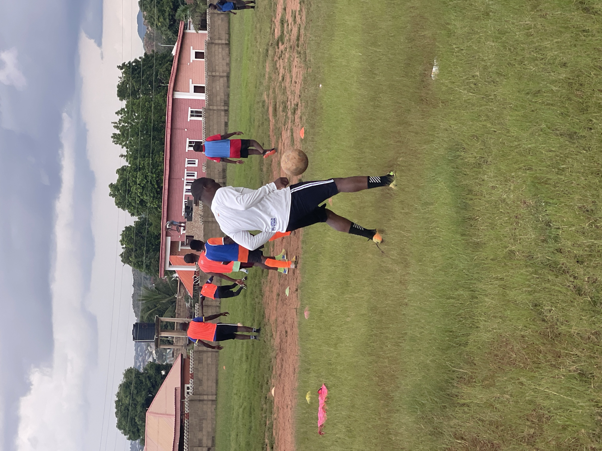 The image depicts an outdoor scene in Jos, Nigeria, featuring a group of young men engaged in a soccer training session. The location appears to be a sports field with patches of grass and dirt. In the background, there are buildings, including a house with a red roof and another with a pink facade, separated by a stone wall. A water tank is visible on a platform behind the buildings, and trees are scattered throughout the landscape. The sky is overcast with dark clouds.
In the foreground, a young man in a white long-sleeved shirt and black shorts with white stripes is actively playing soccer, with the ball at his feet. He is wearing black socks with white stripes and bright yellow soccer cleats. Behind him, other individuals are also present on the field. Some are wearing orange vests over their shirts, and at least one is in a blue vest. They appear to be in various stages of training, with some actively participating in the drills and others observing. Scattered on the field are several small orange and yellow cones, likely used for training exercises. A crumpled pink cloth is visible on the grass to the left of the main subject. The overall atmosphere suggests a focused sports practice.