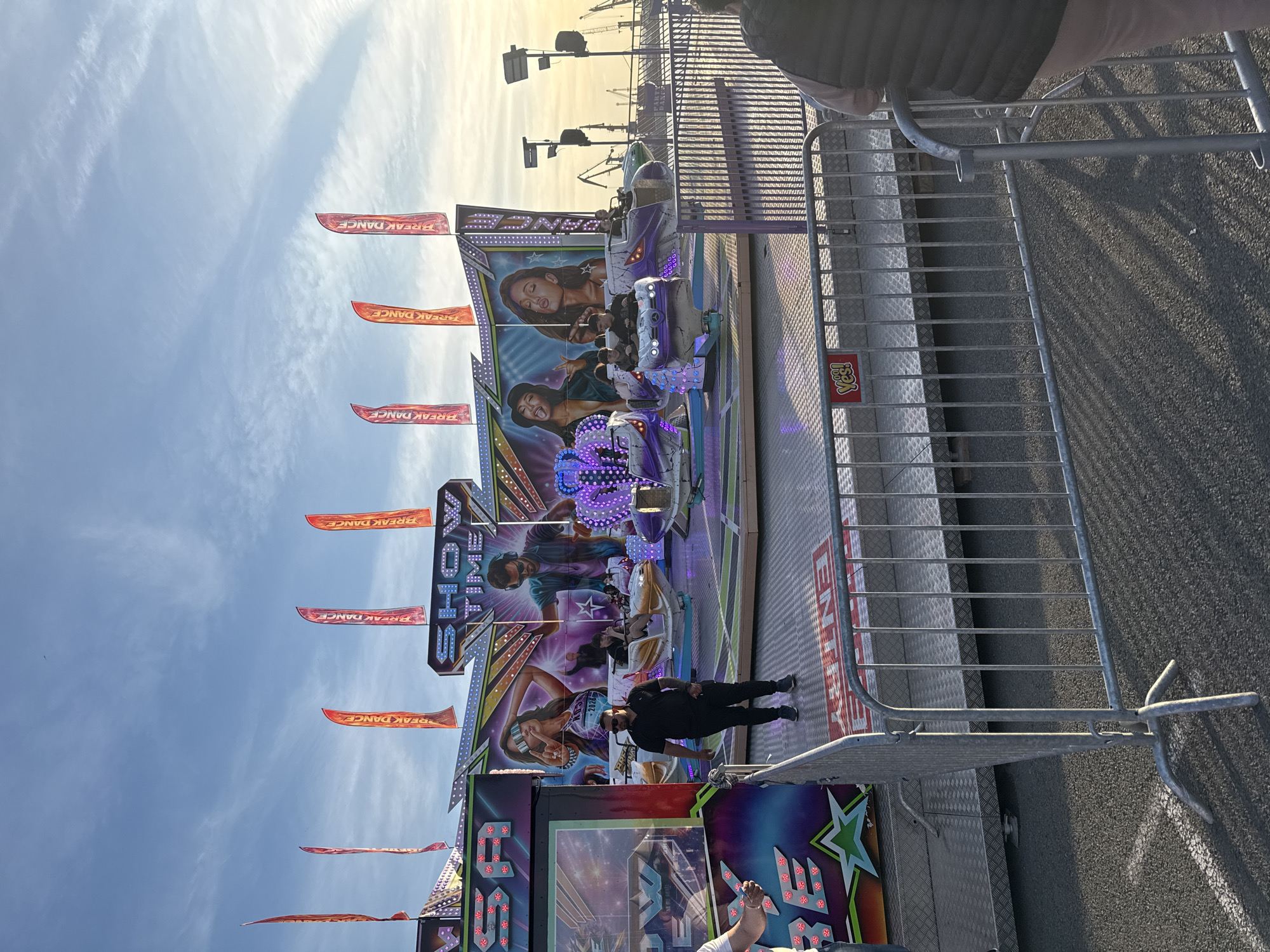 The image captures a vibrant scene at a fairground in Burgas, Bulgaria, likely in 2026 based on signage visible on the ride. The primary subject is a "Break Dance" ride, decorated with colorful graphics and "SHOWTIME" branding. People are riding the attraction. Flags with "Break Dance" printed on them are attached to the ride. A man in a black outfit stands near the ride, possibly an operator. The environment suggests an outdoor amusement park with a partially overcast sky. Metal barriers are in place, likely for crowd control.