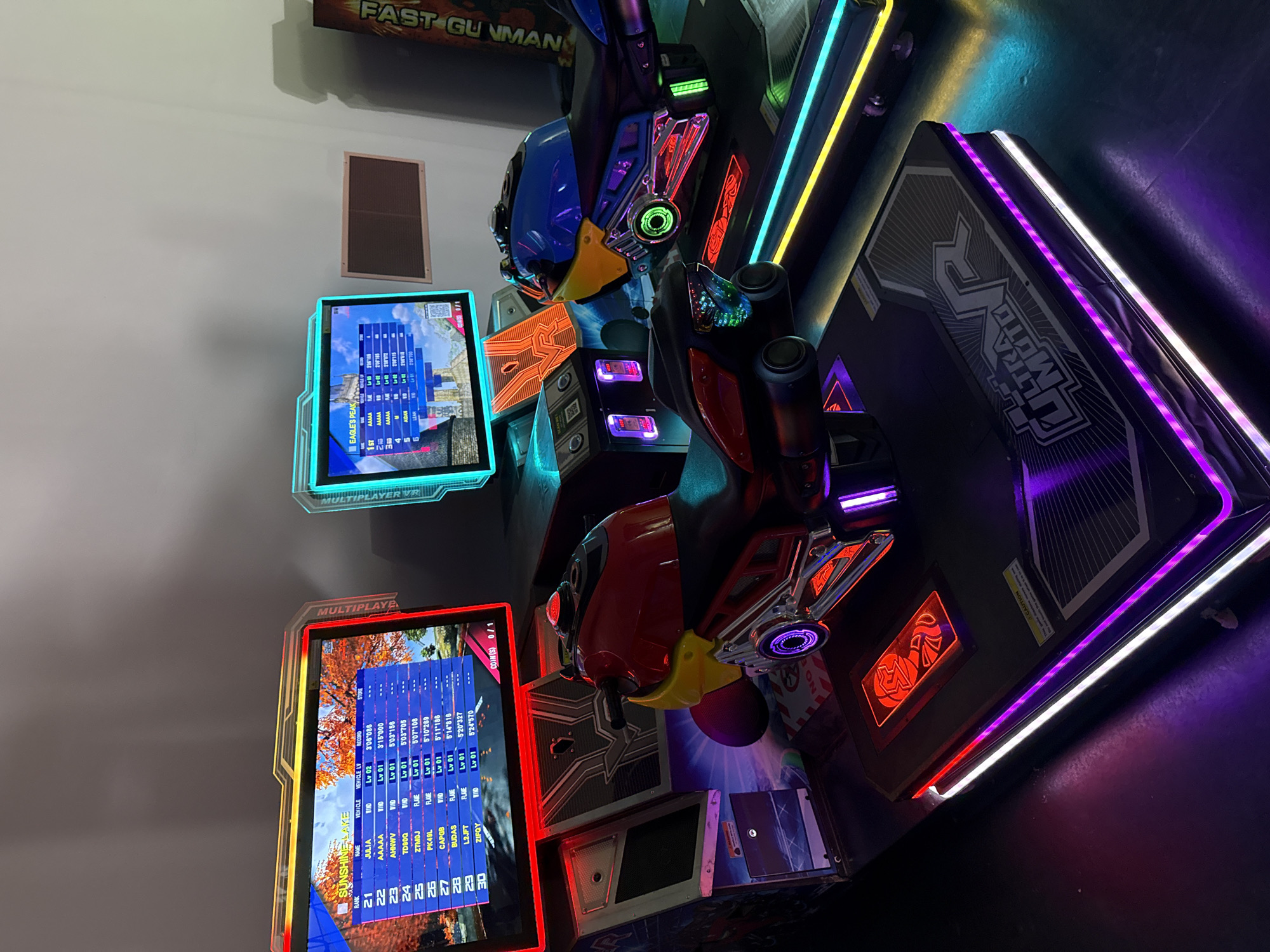 The image shows two arcade racing simulation machines, specifically motorcycle-themed, with a third, partially visible machine to the right. The machines feature illuminated screens displaying game information and leaderboards, as well as brightly lit neon trim around the bases. The motorcycles are brightly colored, with the one on the left being red and yellow, and the one on the right being blue and yellow. Both are designed for a single rider. The machines are placed on a dark floor that reflects the neon lighting. The background is dimly lit, with a plain wall visible behind the arcade machines. A sign with the text "FAST GU..." is partially visible on the right side of the image. The overall environment appears to be an arcade or entertainment center. There are no people visible in the image. The specific location in Bodrum, Turkey, cannot be determined from the visual information provided in the image.