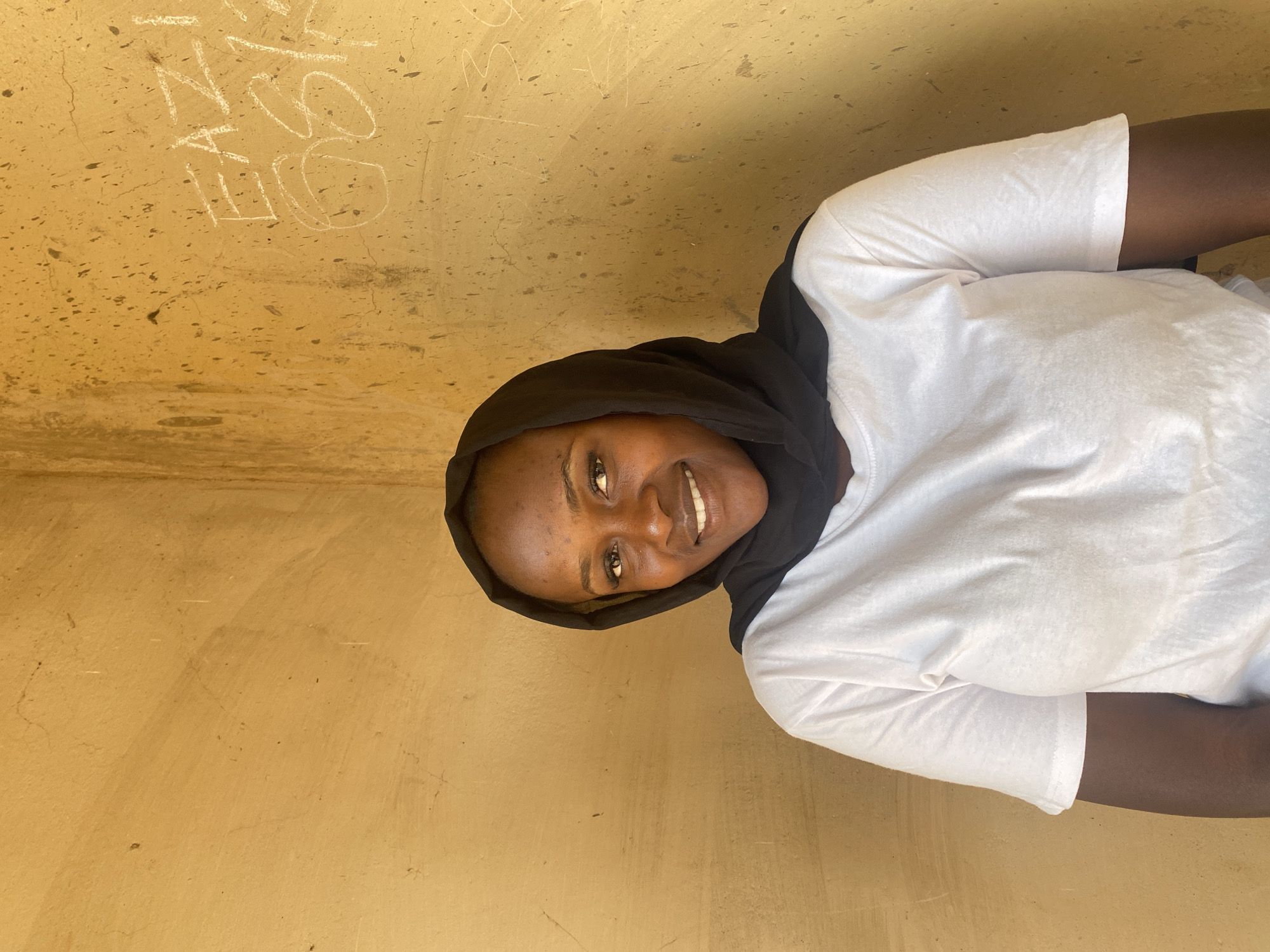 A medium close-up portrait shows a young person, likely female, with dark skin, smiling broadly at the camera. She is wearing a black head covering or hoodie that frames her face, and a white short-sleeved t-shirt. Her expression is cheerful and friendly, with her teeth visible as she smiles.
The background is a textured, light brown or yellow wall, possibly made of concrete or stucco. The wall has a somewhat rough surface with visible cracks, minor indentations, and what appear to be paint splatters or dirt marks. Written on the wall in white chalk or paint are several letters and symbols. Clearly visible are "AN" on the top left, followed by a stylized "T" or "J", and then an "S" or "8" that appears open at the bottom. Below these, there is a circular scribble. Fainter white letters, possibly "M" and "V", are visible further down on the wall. The lighting is bright and even, suggesting it is daytime. The overall setting appears to be an indoor or shaded outdoor area in Kuru Dungus, Nigeria.