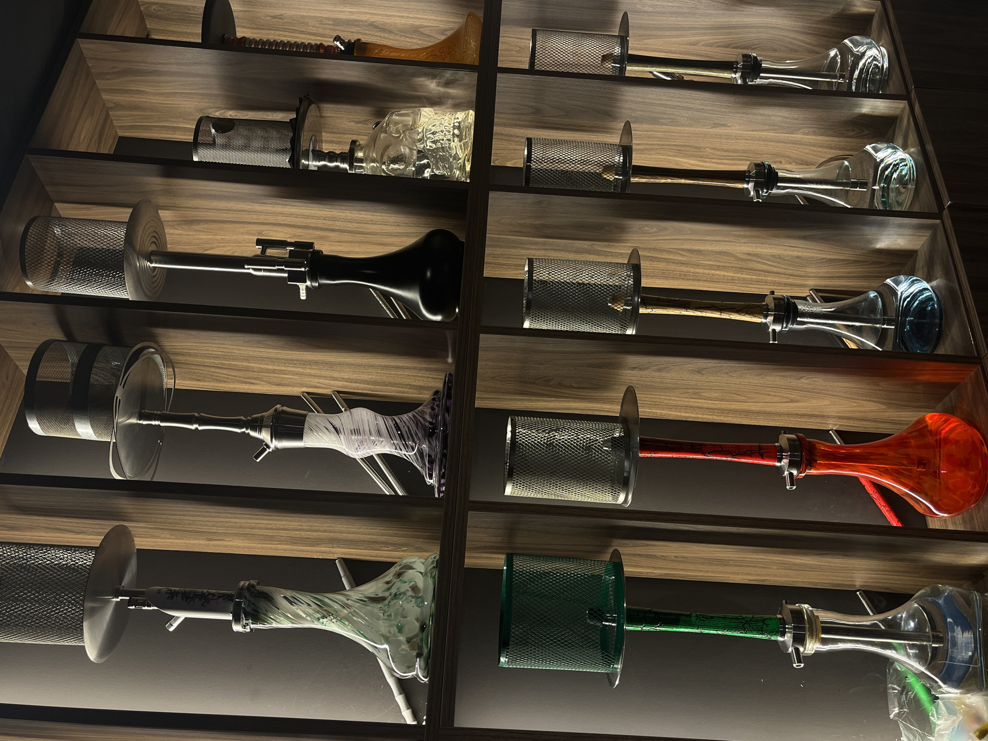 The image shows a wall display of shisha pipes in what appears to be a retail or lounge setting. There are two shelves, each displaying multiple shisha pipes. The top shelf features four shisha pipes with decorative glass bases in swirling green, purple, and clear colors, as well as a black one and one with a clear skull-shaped base filled with liquid. The bottom shelf displays six shisha pipes, with bases colored green, red, blue, and clear, all filled with liquid. The pipes are arranged vertically in separate compartments formed by wooden panels. The lighting is dim, highlighting the shisha pipes. The location is Plovdiv, Bulgaria.