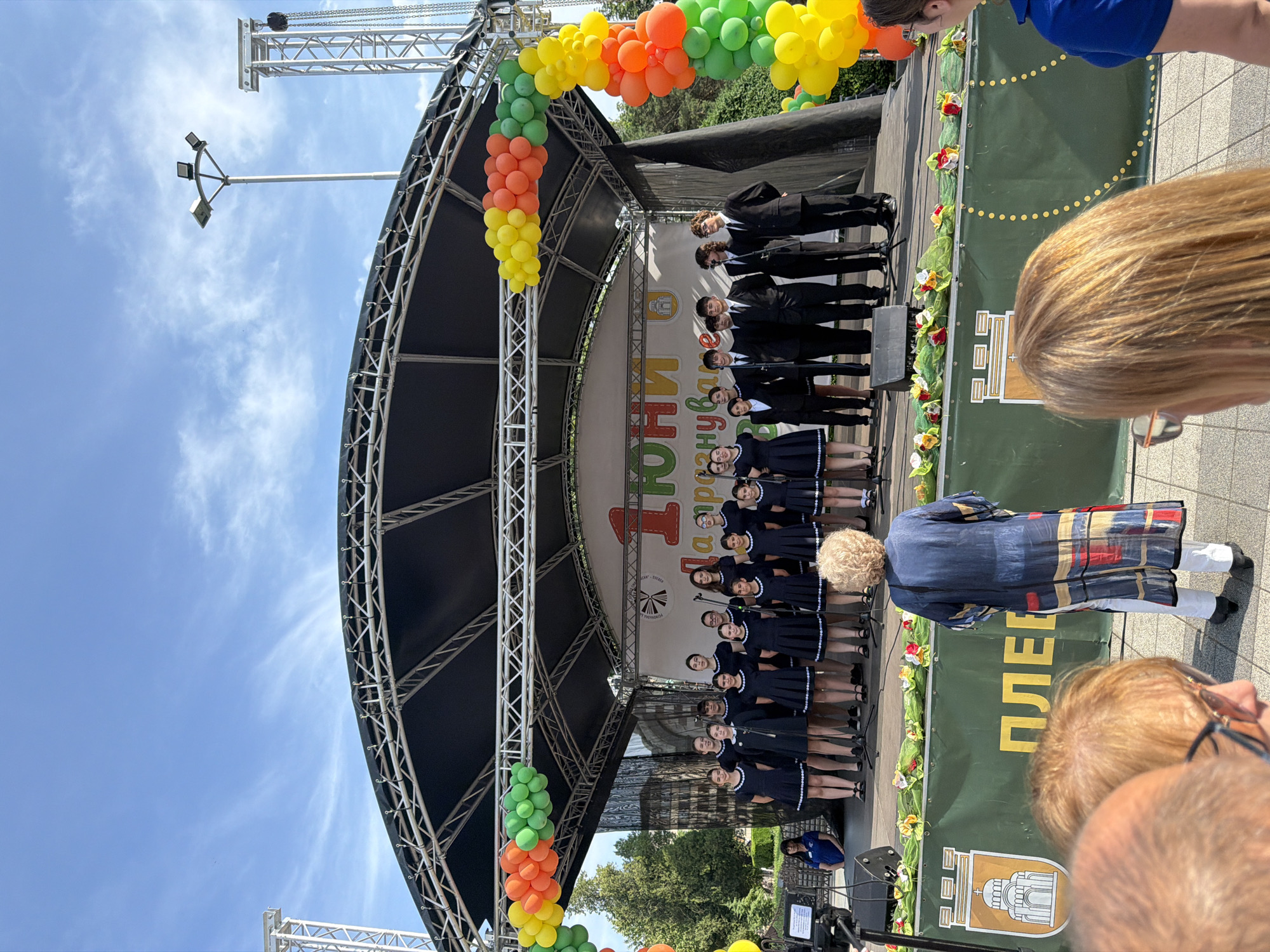 The image shows a choral group performing on a stage in what appears to be the city of Pleven, Bulgaria. The stage is adorned with balloons in orange, green, and yellow. A banner with the name "Pleven" in Cyrillic is visible behind the performers. The choir consists of young individuals, some wearing navy blue dresses with white trim and others wearing suits. An audience watches the performance. The sky is blue.