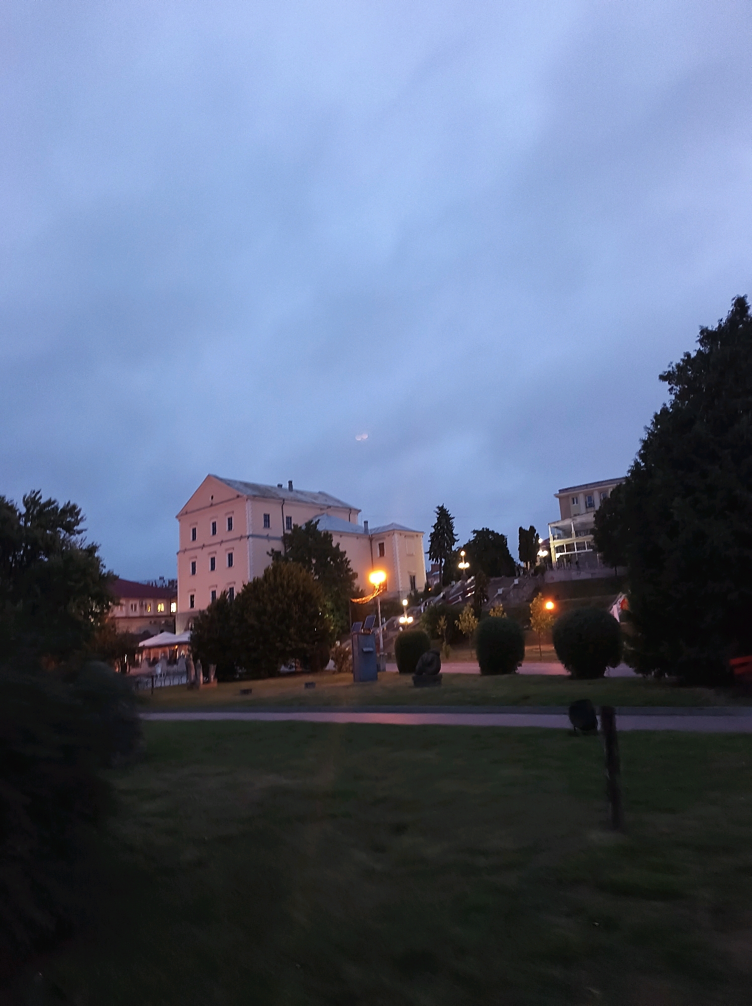 The image depicts a twilight scene in Ternopil, Ukraine, featuring several buildings and a grassy area. In the background, a large, pale yellow building with multiple windows and a sloped roof dominates the left side of the frame. To its right, another building with a similar architectural style and balconies is visible. Further to the right, a modern building with balconies and white railings stands against the darkening sky.
The foreground consists of a well-maintained grassy lawn with scattered bushes and trees. Several lampposts are illuminated, casting a warm glow on the surroundings. There is a suggestion of a pathway or road running through the grass. In the far distance, a faint, round shape in the sky might be the moon or a reflection. No people are clearly visible in the image. The overall atmosphere is calm and serene, typical of a late evening in a city park or a residential area.