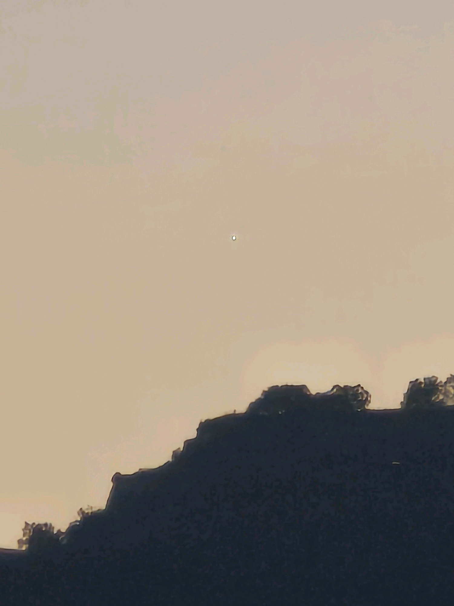 A distant, bright white circular object is visible in the upper center of the frame against a pale orange sky. Below it, a dark silhouette of a hilly, tree-covered landscape dominates the lower portion of the image. The terrain is uneven, with the tops of trees appearing jagged against the sky. The overall impression is of a view from a distance, likely at dusk or dawn, with the bright object potentially being a star, planet, or distant light. The specific location in Karachayevsk, Russia, is not visually identifiable within the image itself, as the scene depicts a natural landscape.