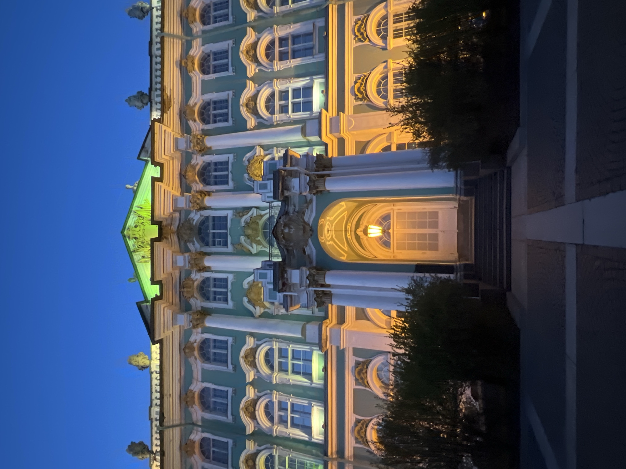 The Winter Palace in Saint Petersburg, Russia, is visible from an exterior, low-angle perspective, set against a darkening blue sky. The architectural details are highlighted with light on the building's facade. The building's columns, windows, and arched structures feature ornate architectural embellishments. The entryway shows illumination from within. Shrubs or trees are partially visible on either side, encroaching into the view of the building, as well as the path to the entrance of the building. A path of pavement leads towards the building, providing a foreground element.
