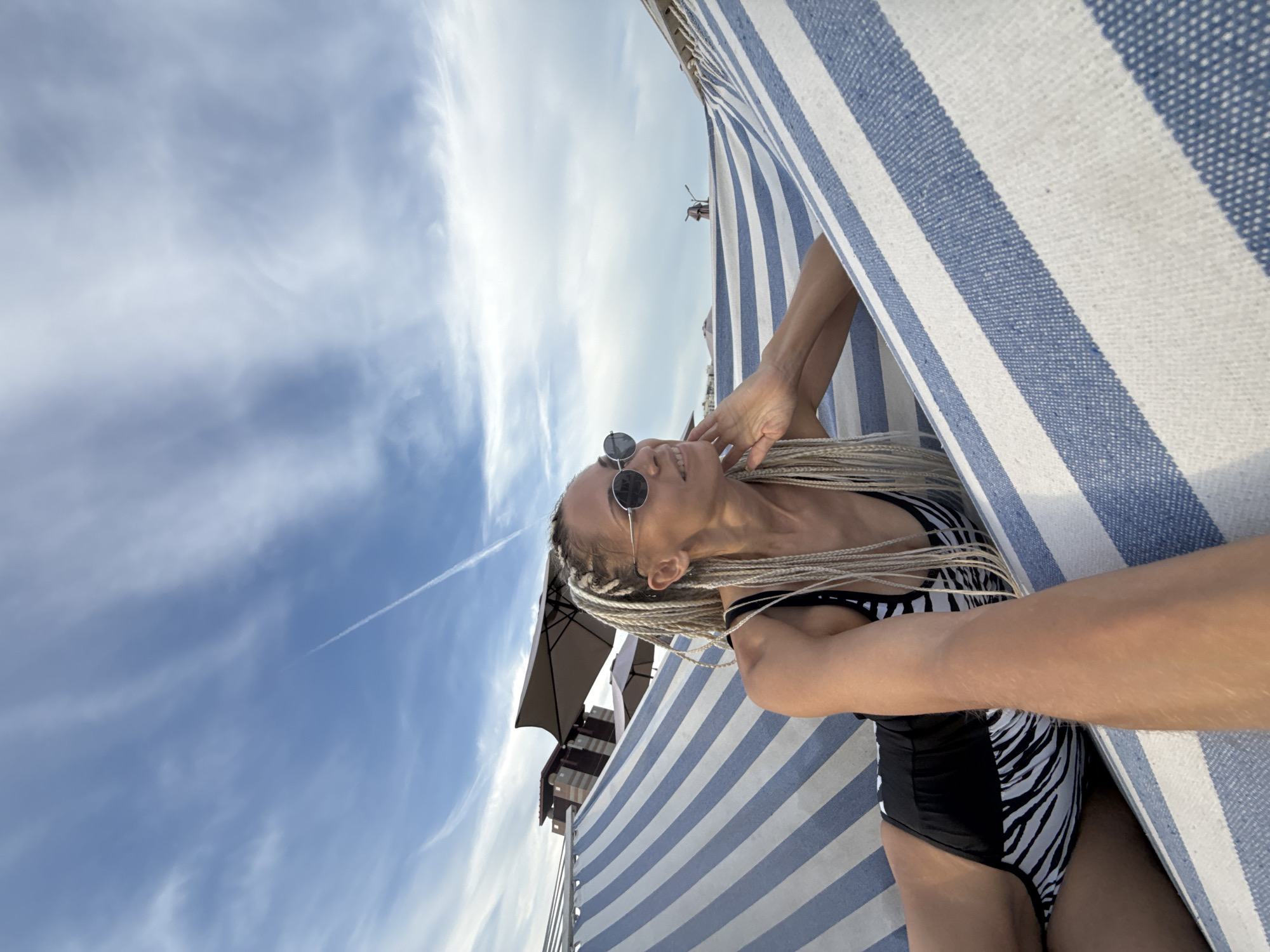A woman with blonde braided hair and sunglasses lies in a hammock with blue and white stripes, wearing a black and white zebra-print swimsuit. She is looking to the right with her hand resting on her chin. The hammock is suspended outdoors, with a clear blue sky and wispy clouds in the background. In the distance, there are some buildings and umbrellas, suggesting a resort or beach area. The scene is calm and relaxed. This image was taken in the city of Rusanovo, Russia.