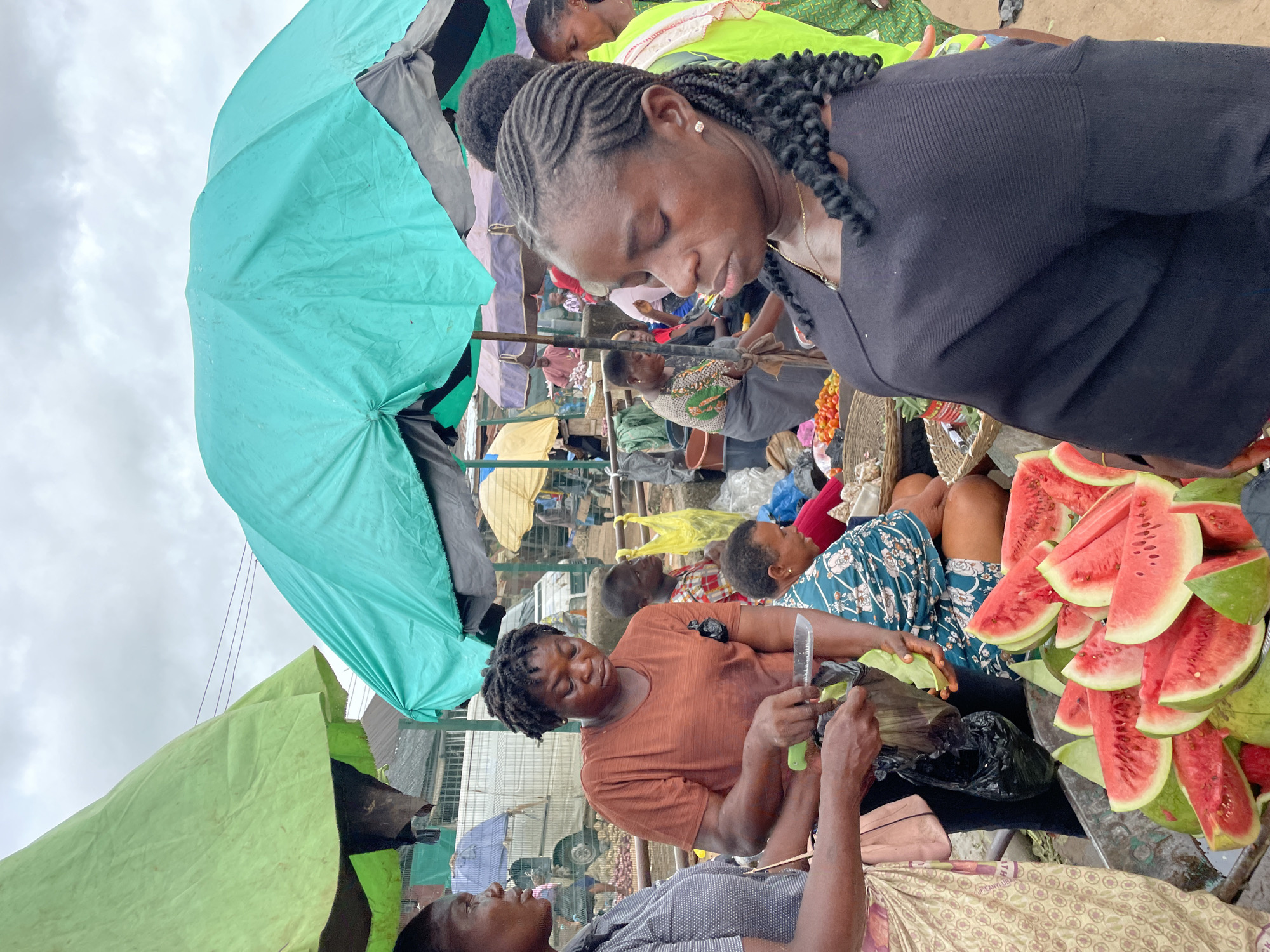 This is a medium shot of a bustling outdoor market in Unknown, Unknown, under a cloudy sky. Several women are gathered around a stall selling watermelons and other produce. In the foreground, a woman in a black top with braided hair looks down, her profile visible. To her left, another woman in a rust-colored t-shirt is slicing fruit, holding a knife in her right hand and a black plastic bag in her left. A pile of watermelon slices is in the lower left and center of the image. Behind them, other market-goers are visible, including a woman in a blue and white patterned top and a boy in a plaid shirt. Various umbrellas and stalls create a colorful backdrop. The ground appears to be dirt.