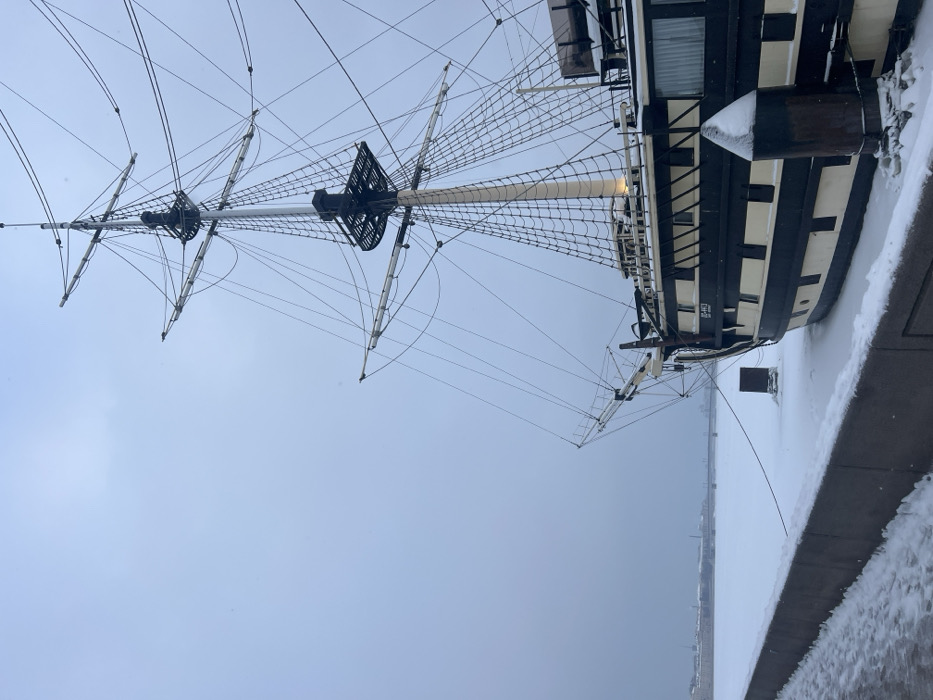 The image captures the starboard bow and foremast sections of a large, multi-masted sailing ship, identified by the visible name "ПОЛТАВА" (Poltava) on its black and off-white striped hull. The ship is moored on the Neva River embankment in Saint Petersburg, Russia, during winter.
The scene is dominated by cold conditions, with snow covering the top of a dark grey concrete embankment in the foreground, the entire surface of the frozen river to the left of the ship, and portions of the ship's deck and mooring bollards. The ship's intricate rigging, including multiple masts, spars, and a complex network of ropes, is visible against a uniformly overcast, light grey sky. In the distant background, across the frozen river, a blurred cityscape with buildings and a bridge is discernible. No individuals are present in the image.
