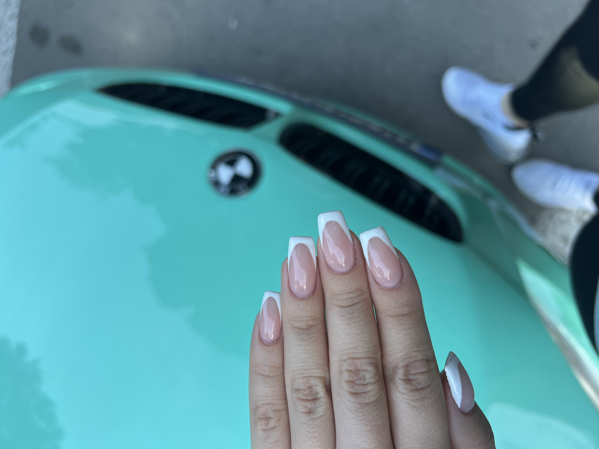 An overhead photograph captures a human hand with manicured nails positioned over the hood of a bright teal BMW vehicle in Asenovgrad, Bulgaria. The hand, displaying a light-to-medium skin tone, features five visible fingernails (thumb, index, middle, ring, pinky), all shaped in a square/coffin style with rounded corners. Each nail is painted with a classic French manicure, comprising a light pinkish-nude base coat and distinct opaque white tips. The car's hood, featuring a visible BMW emblem and part of its signature kidney grille, reflects soft, diffuse light from above. In the lower right quadrant of the image, blurred lower legs and feet of a person wearing black leggings and white athletic sneakers are visible, standing on a textured gray paved surface.