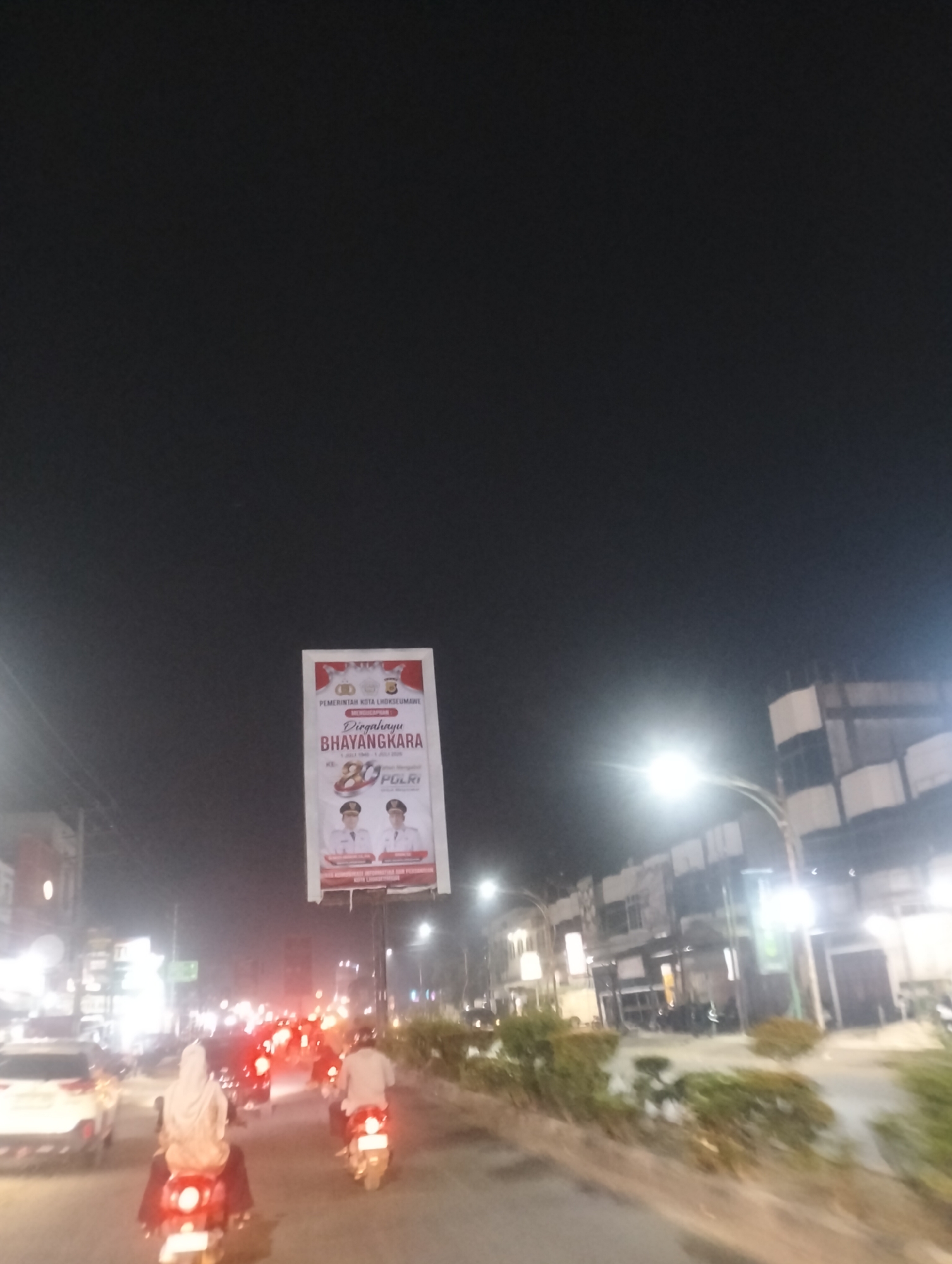 This is a vertical, nighttime, outdoor shot of a street scene in Lhokseumawe, Indonesia. The scene is illuminated by streetlights and the lights from passing vehicles and buildings. The sky is dark, suggesting it is late at night.
The main subject is a large billboard erected on a pole. The billboard features text and images. At the top, it reads "PEMERINTAH KOTA LHOKSEUMAWE" (Government of Lhokseumawe City). Below this, it says "Dirgahayu BHAYANGKARA" (Happy Anniversary BHAYANGKARA), with "KE-77" (77th) and "1 JULI 1946 - 1 JULI 2023" indicating the date range. There are images of two men in formal attire, likely officials, and the POLRI logo.
In the foreground, several motorcycles are visible, suggesting traffic is moving on the road. One motorcycle in the center has a rider wearing a hijab, and another rider is visible further back. Other vehicles, including cars, are also present in the traffic. Buildings line the street on the right side of the image, some with illuminated windows. The overall impression is a busy urban street at night.