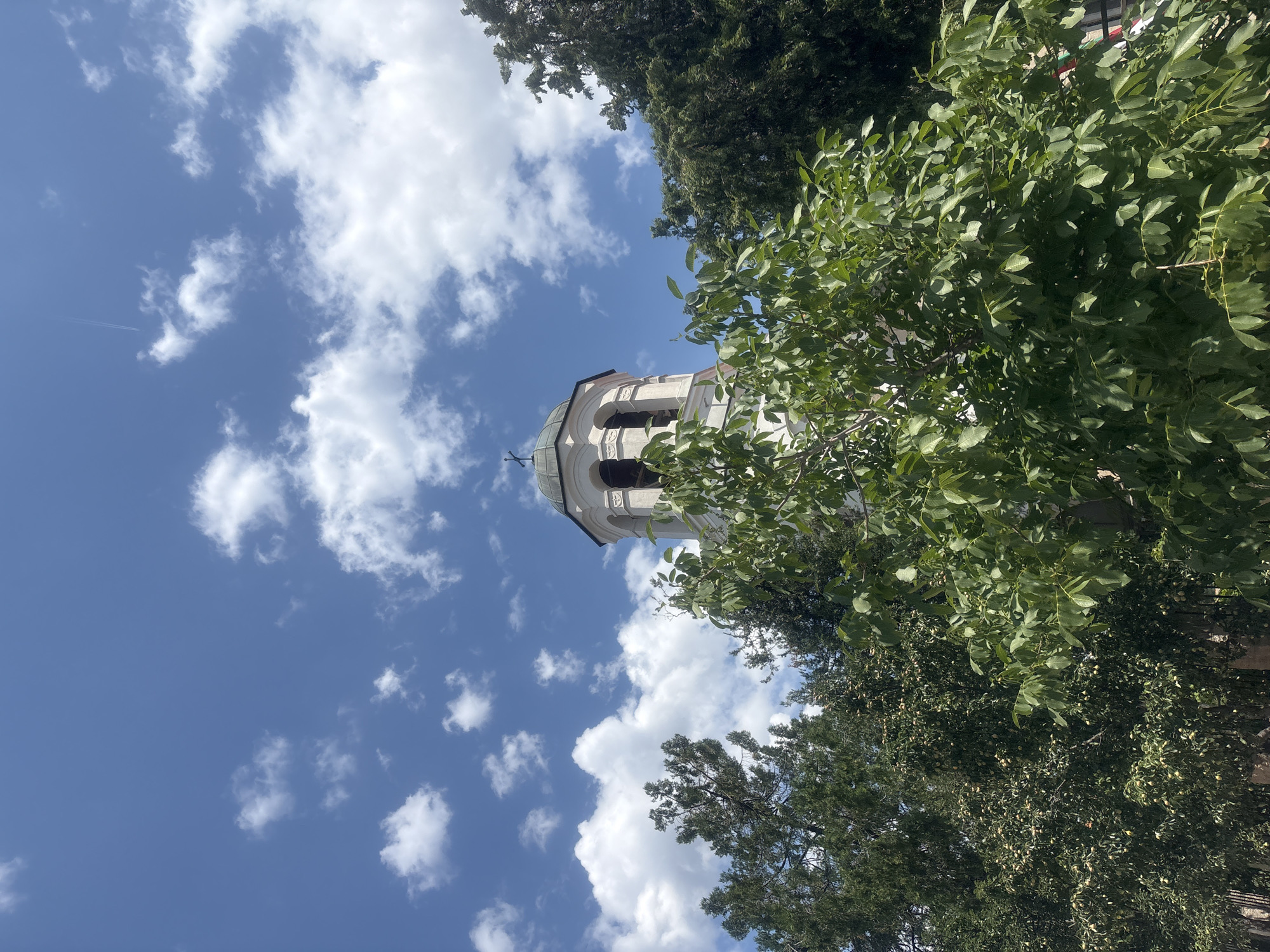 The image captures a view of a white bell tower topped with a cross, partially obscured by lush green trees. The bell tower has arched openings and a domed roof. Above, a bright blue sky is dotted with fluffy white clouds, and a faint airplane contrail can be seen. The trees in the foreground and midground are dense with foliage, suggesting a warm, sunny day. The scene appears to be set in Strelcha, Bulgaria, possibly depicting a church or a historical landmark surrounded by nature. No people are visible in the frame.