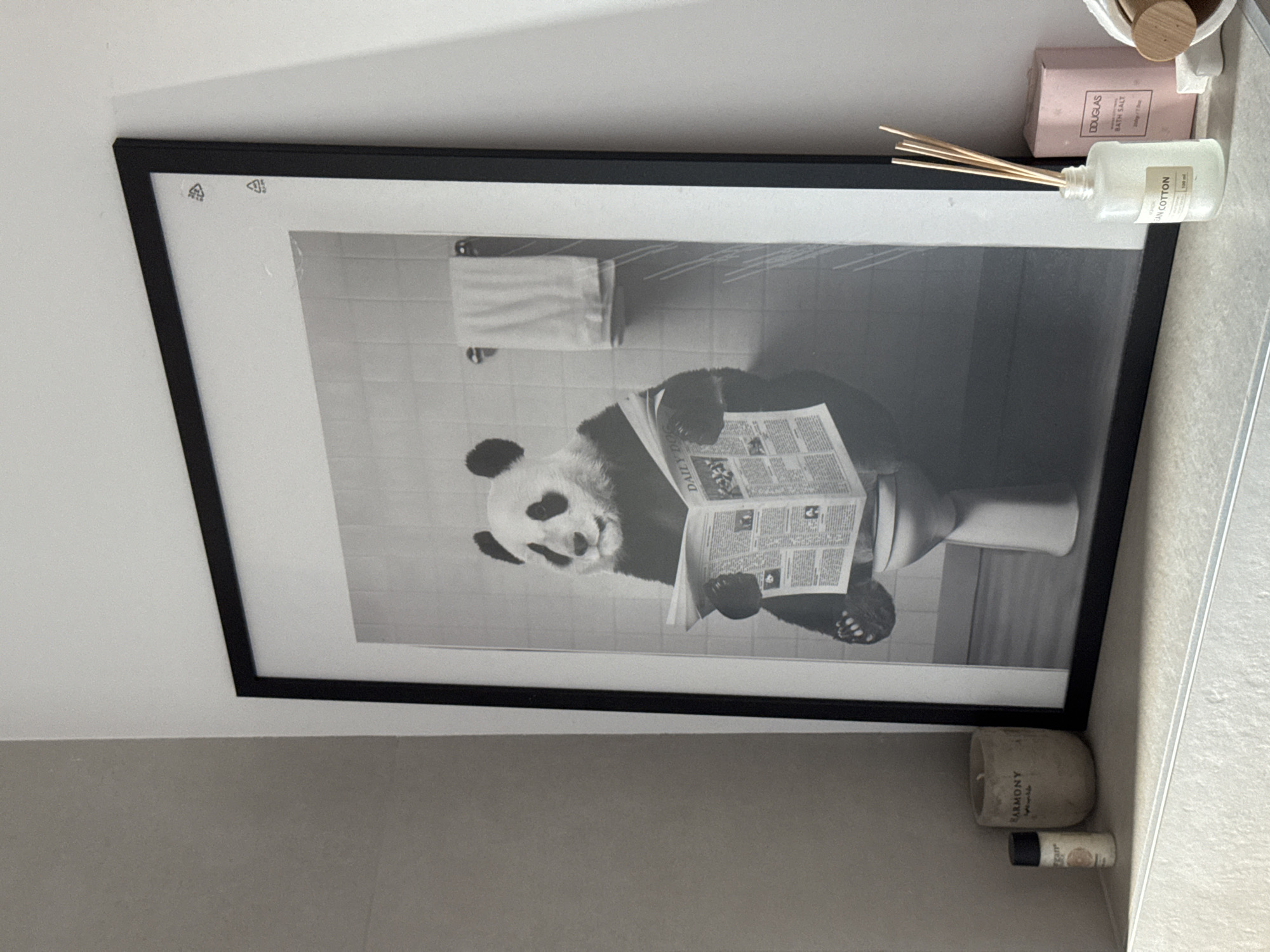 A black-framed picture of a panda reading a newspaper while sitting on a toilet is mounted on a wall in an indoor bathroom. The picture is in black and white. Next to the picture are various items, including a bottle with wooden sticks, a pink box, and a stone-like container. The environment consists of tiles and plain walls. The location is Unknown, Unknown. There are no people present in the image.