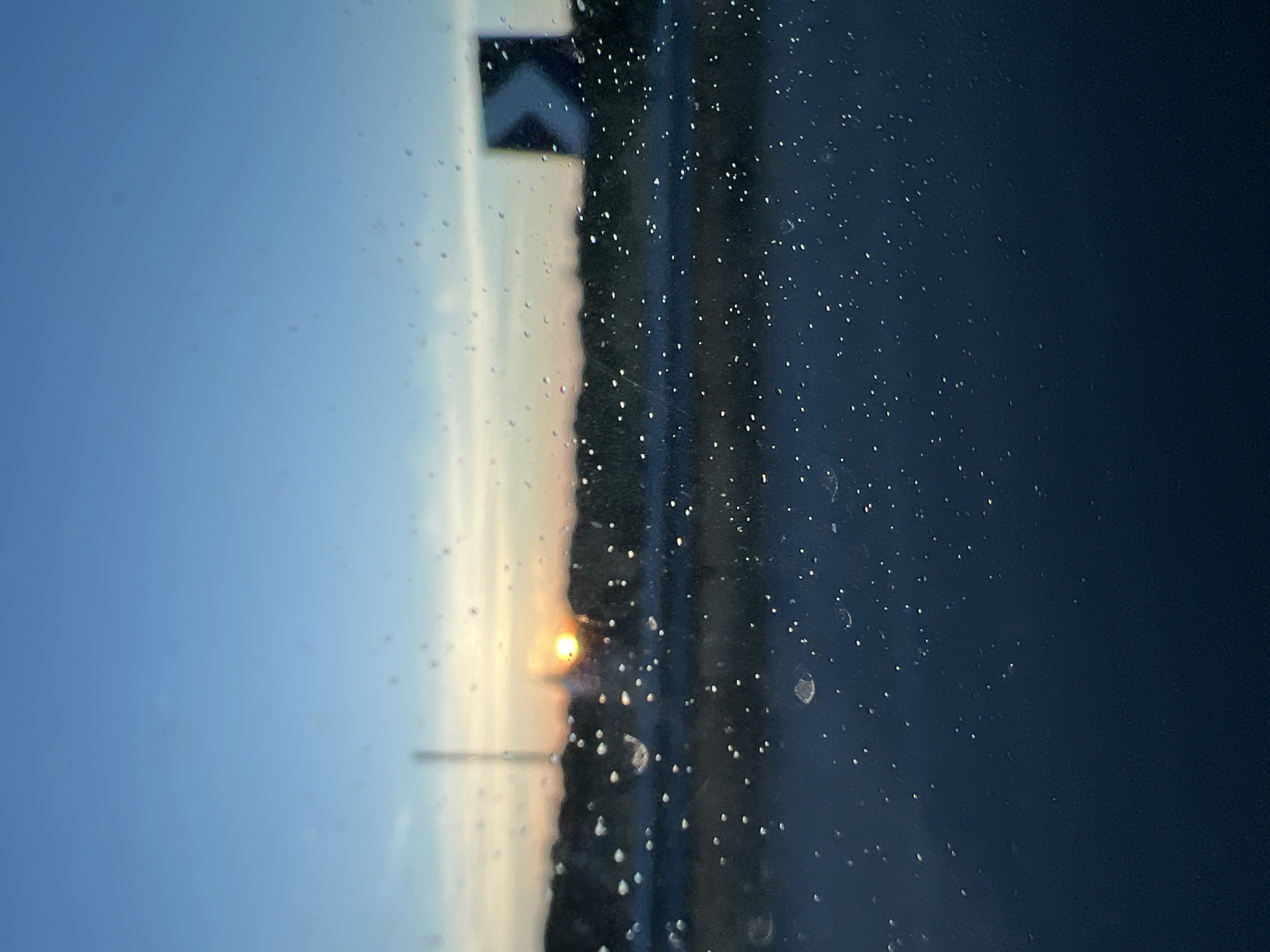 This image, captured in Cassano delle Murge, Italy, depicts an outdoor scene at either dawn or dusk, viewed through a transparent surface heavily covered with water droplets. The foreground is dominated by these sharply focused, spherical water droplets of various sizes on a window, creating a bokeh effect on the background elements.
The midground reveals a highly blurred landscape. A dark, undulating horizon line, likely composed of distant trees or hills, spans the width of the frame. A bright, intense orange-yellow orb, representing the sun, is positioned centrally on this horizon, partially obscured by the silhouetted terrain. To the left of the sun, a faint, tall, slender vertical structure, possibly a pole or mast, rises against the sky. To the right, a dark, blurred rectangular object with a prominent white chevron pointing left is visible, consistent with a road sign. Below the horizon line, a dark, elongated band suggests a road surface or barrier.
The background consists of the sky, which transitions from a light orange-yellow hue directly above the sun, indicative of twilight, to a clear, progressively deeper blue in the upper portion of the image. No distinct cloud formations are discernible. There are no people or explicit actions depicted. The scene suggests observation from a stationary or moving vehicle.
