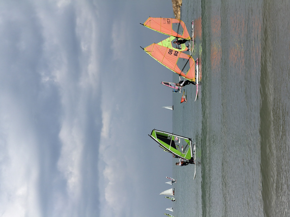 The image displays a maritime scene depicting multiple windsurfers on the Mediterranean Sea, off the coast of Tel-Aviv, Israel. The sky is overcast with layered grey clouds dominating the upper two-thirds of the frame. In the foreground, three individuals, each wearing a dark wetsuit, are actively windsurfing. The leftmost individual stands on a white board, managing a green and black sail marked with the number '113'. The central individual also stands on a white board, controlling a peach-orange sail marked 'OII 110'. The rightmost individual stands on a white board, operating a similar peach-orange sail marked '157'. All three subjects are positioned facing towards the right side of the frame. The water surface shows small, visible wave formations. In the mid-ground and background, several additional windsurfers with sails of various colors (including pink and white) and at least three traditional sailboats with white sails are visible. A rocky landmass is present along the distant right horizon.