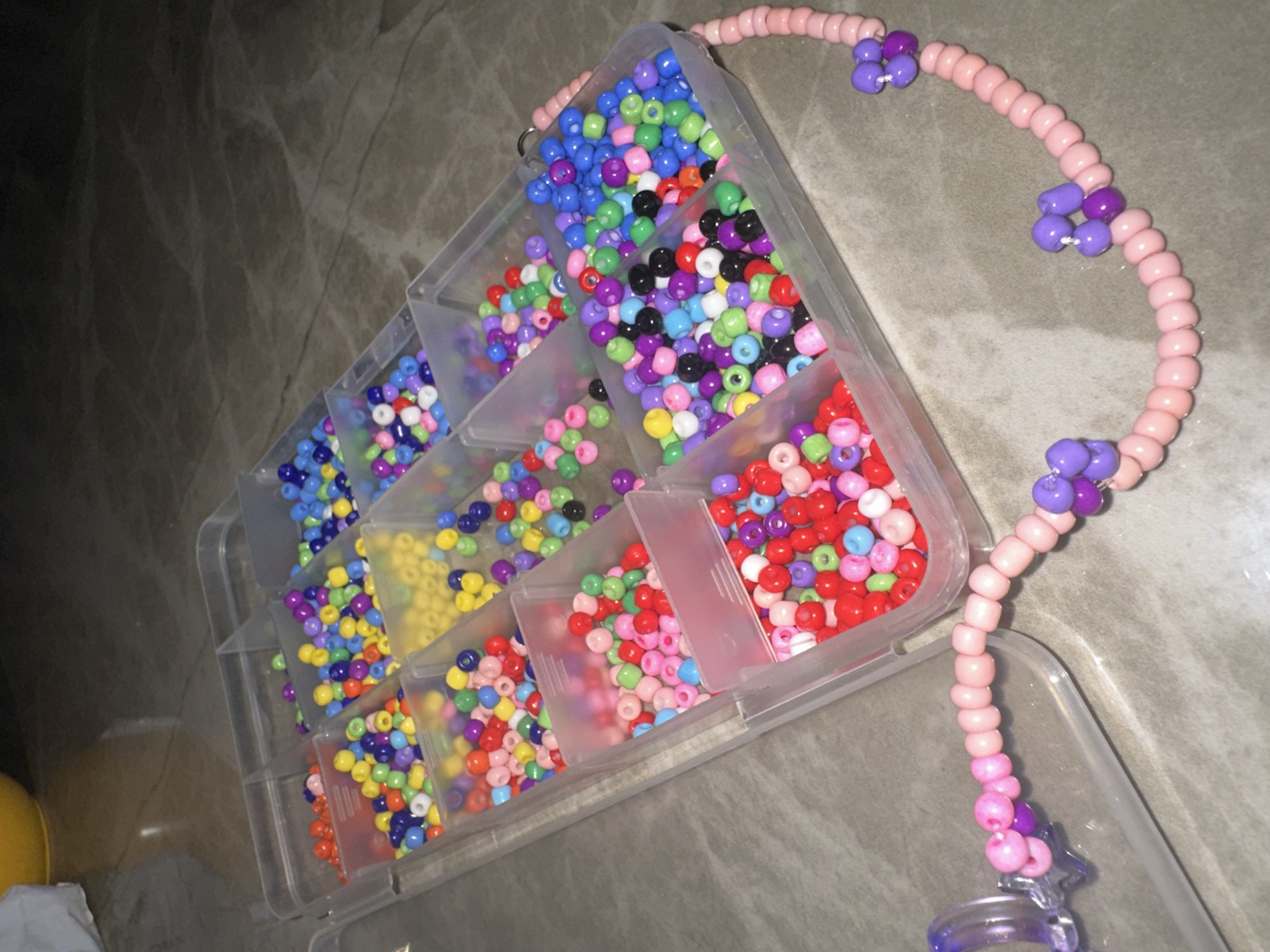 The image shows a clear plastic container with multiple compartments filled with colorful plastic beads. The beads vary in color, including blue, green, yellow, red, pink, purple, black, and white. An unfinished bead necklace made of light pink beads with clusters of purple beads is lying next to the container. The background appears to be a dark surface, potentially a countertop or floor. The location in Bac Nha Trang Ward, Vietnam, is not determinable from the given visual information.